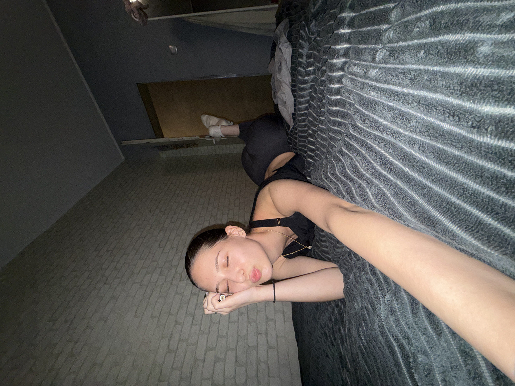 A woman is lying on a bed, apparently indoors. The woman is young and is wearing a black sports bra and black leggings. She is also wearing a black bracelet. She has a necklace. She is lying on her side with her eyes closed, on a gray, textured blanket or comforter. Her head is near a gray brick wall. Her feet are bare and she is wearing white socks. There is a brown cabinet or shelf on the back wall. The picture is taken at a tilted angle. There is no indication of the location being in Sofia, Bulgaria.