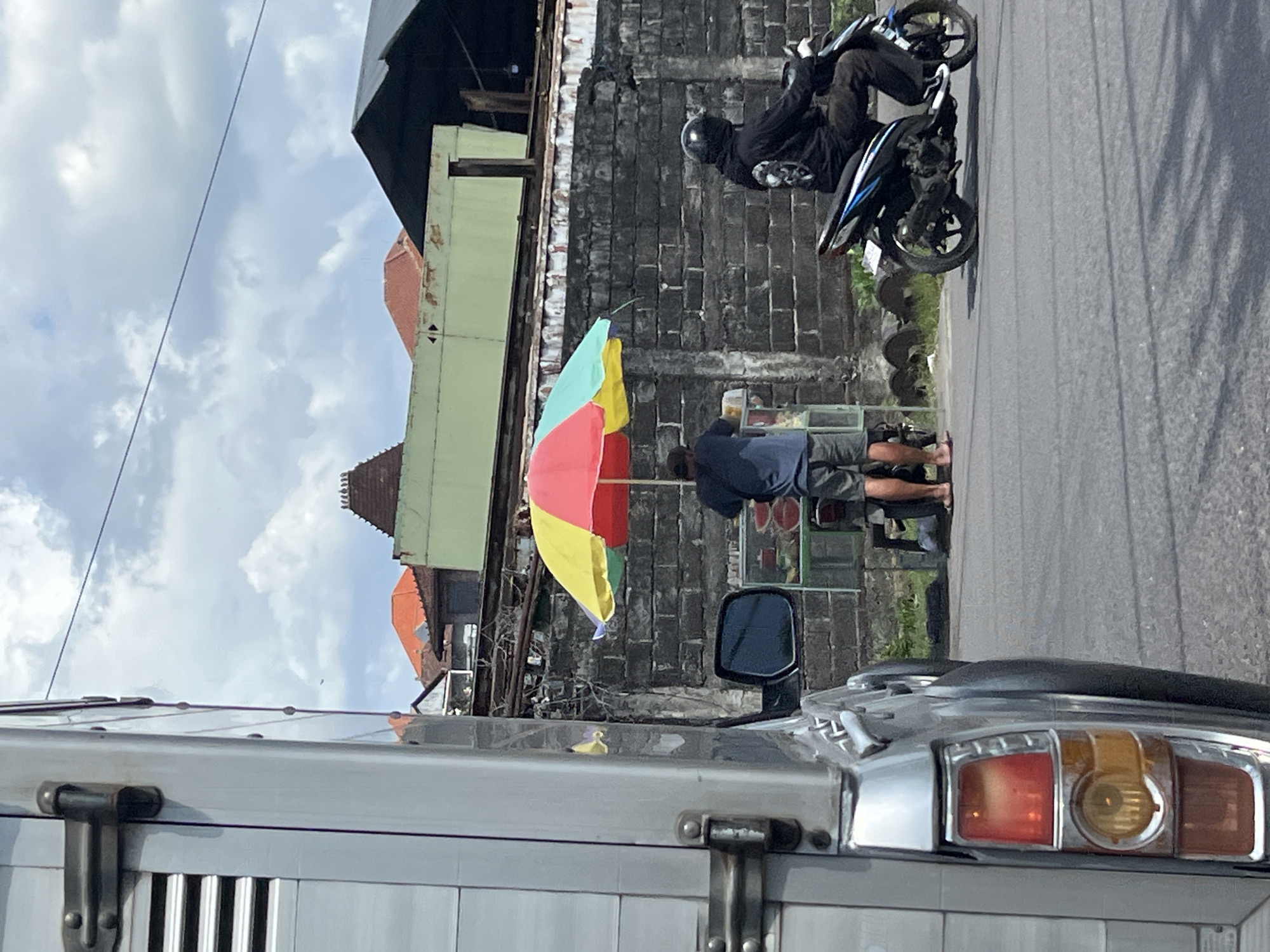 A street vendor with a multi-colored umbrella stands by a cart filled with fruits, likely coconuts and watermelons, in Denpasar, Indonesia. The vendor is dressed in a blue t-shirt and shorts. To the right, a person wearing a black hoodie and a helmet rides a black and blue motorcycle. The scene is set against a textured brick wall and a backdrop of buildings with orange and green roofs. In the foreground, a silver vehicle is partially visible on the left. The sky is partly cloudy, with a few trees and electrical wires visible.