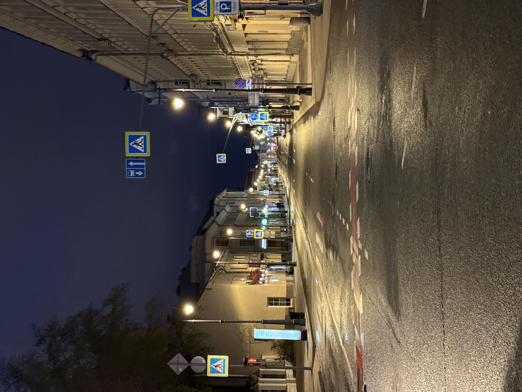 The image depicts a street scene in Moscow, Russia, at night. The street is wet and reflects the light from the street lamps. The buildings on either side of the street are illuminated. Several traffic signs are visible along the street. There are no people visible in the frame. The environment suggests a calm and quiet urban night.