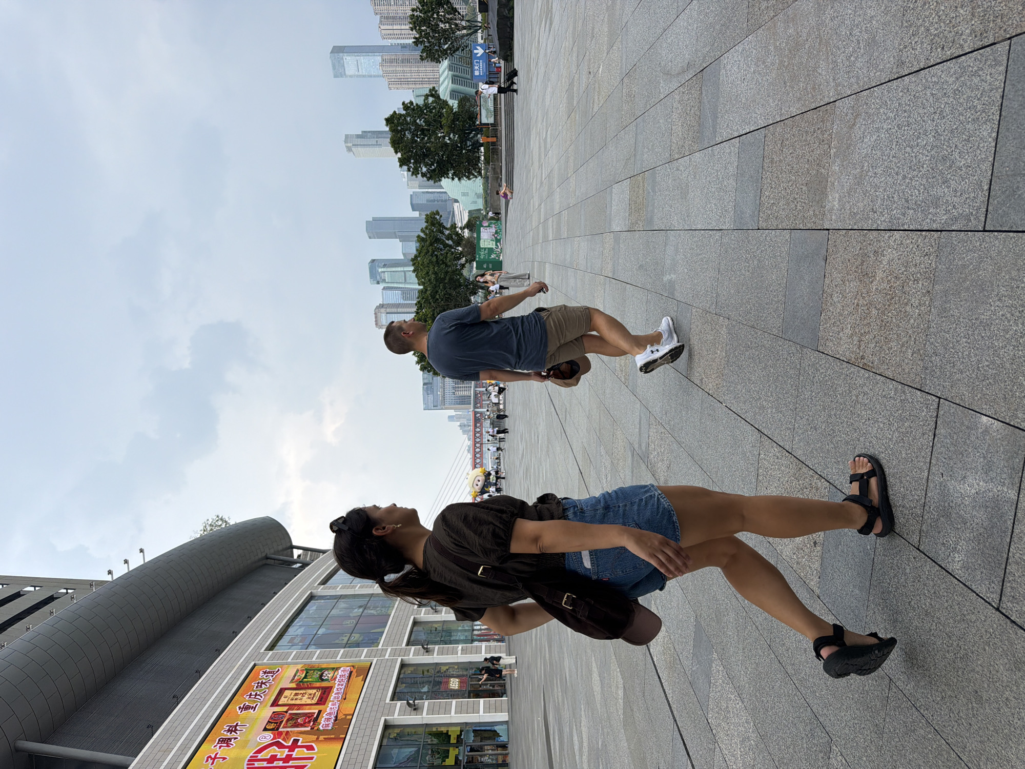 The image captures an outdoor scene in a modern urban environment, likely in the Yuzhong District of Chongqing, China, given the architectural style and cityscape. In the foreground, a woman with her hair in a ponytail is walking to the left, wearing a dark short-sleeved shirt, denim shorts, and black sandals. A brown bag is slung over her right shoulder, with a brown baseball cap peeking out from it. To her right, a man is walking towards the right, wearing a dark blue t-shirt, khaki shorts, and white athletic shoes. He is holding a brown baseball cap in his right hand.
The ground is paved with large, gray rectangular tiles. Behind the individuals, a modern building with a curved facade and large windows is visible on the left, featuring a brightly lit digital advertisement. In the background, a cluster of tall skyscrapers dominates the skyline under a cloudy sky. Further in the distance, a suspension bridge can be seen. Several other people are scattered throughout the plaza, engaged in various activities such as walking and standing. The overall impression is one of a bustling public space within a contemporary Chinese city.