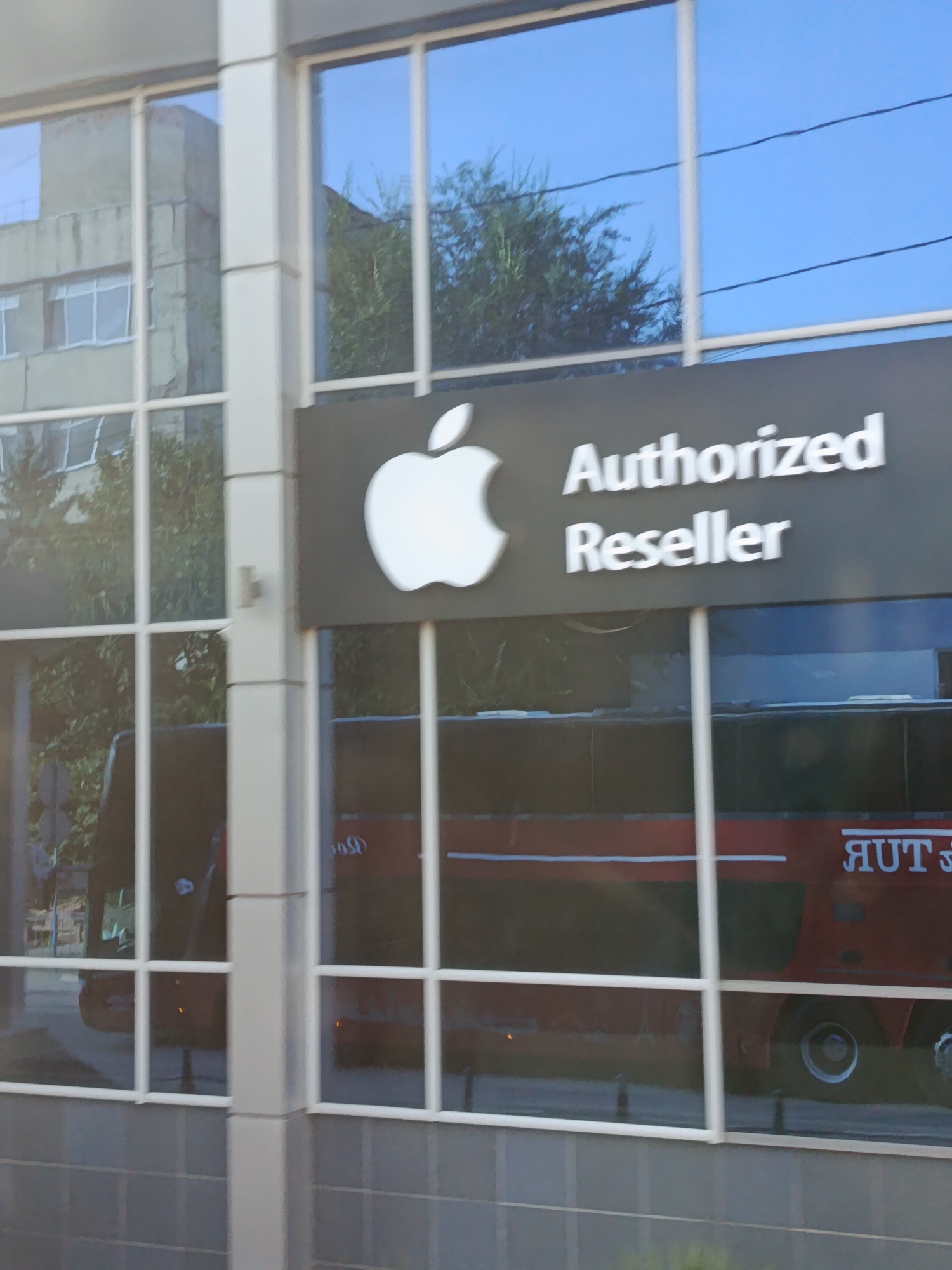 An exterior shot of an Apple Authorized Reseller store in Cahul, Moldova, features a prominent black sign with a white Apple logo and the text "Authorized Reseller." The store has large, reflective glass windows that show reflections of trees, the sky, and a red bus with text written in Cyrillic script. The building's facade is a mix of grey concrete and white vertical elements. The lower portion of the building is covered in grey tiled blocks.