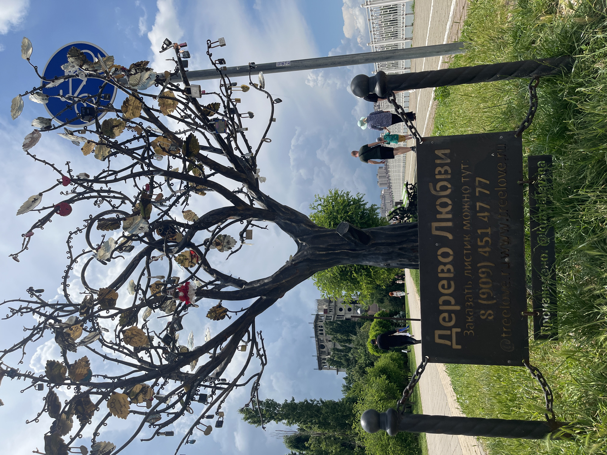 In Novaya Adygeya, Russia, a metal tree sculpture adorned with numerous padlocks and metal leaves stands in a park. A dark metal sign in front of the sculpture reads "Дерево Любви" (Tree of Love), followed by a phone number and website address. In the background, a paved walkway runs alongside a white railing, with people walking on it. Trees and a partially constructed building are visible behind the walkway. The sky is partly cloudy.