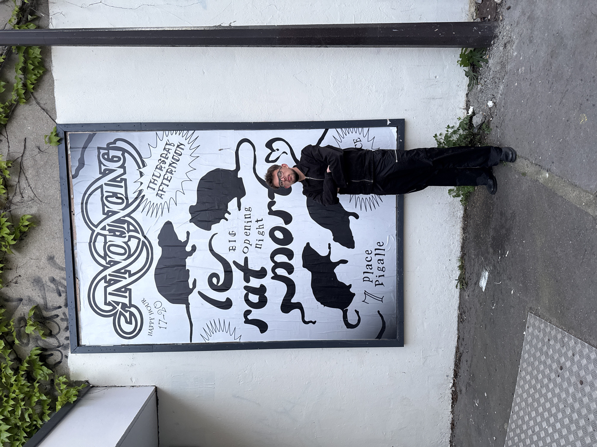 A young man stands beside a large, black-framed poster affixed to a white wall in an urban setting. The man, appearing to be a young adult, wears glasses, a black jacket, and black trousers, striking a pose with crossed arms and a subtle pout. To the left of the poster, ivy cascades down a concrete surface, while a grey sidewalk with patches of weeds and a metal grate extends to the right. A dark, horizontal beam or pipe runs along the top of the wall.
The poster itself is a black and white design, showing some surface crumpling. It prominently features several stylized silhouettes of rats. Various texts are visible: "ANNOUNCING" (partially visible), "let rat morty" (likely the name of an event or venue, in a large, distinctive font), "BIG opening night," "THURSDAY AFTERNOON," and "HAPPY HOUR 17-20." Crucially, it lists "7 Place Pigalle" as the location. This detail, combined with the provided context, indicates the scene is in Paris, France. The daytime lighting, without harsh shadows, suggests an overcast day. The poster promotes an upcoming event, likely an opening with a happy hour, while the man's pose adds a casual, perhaps promotional, element to the scene.