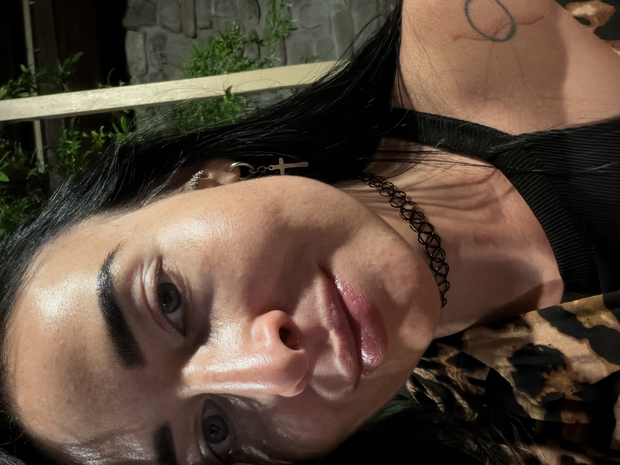 This is a close-up, eye-level, indoor portrait of a woman in Sochi, Russia. She has dark hair, dark eyebrows, and is wearing a leopard print top and a black choker. She has a cross earring in her left ear and a tattoo on her right arm. The lighting suggests artificial indoor lighting, possibly with some natural light filtering in, creating highlights on her skin. The background appears to be a stone wall and some greenery, possibly part of an outdoor seating area or a restaurant in Sochi.