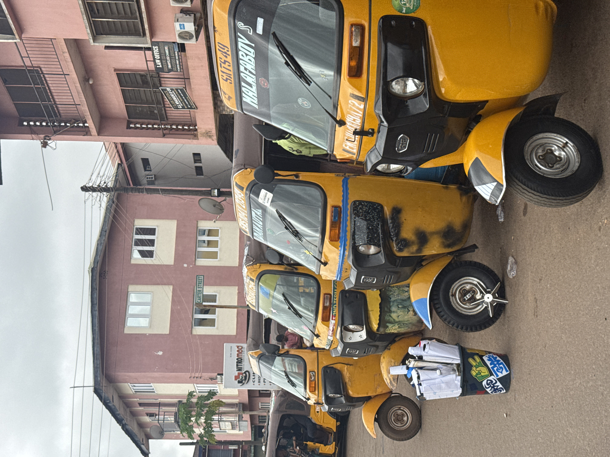 The image shows a street scene in Enugu, Nigeria, featuring a row of yellow auto-rickshaws parked along a road. The auto-rickshaws are distinctive three-wheeled vehicles with covered passenger compartments. Several of them have names or numbers painted on their windshields, such as "MALAFABOY S" and "SATS 49". In the foreground, a black bucket is filled with white umbrellas and other items, decorated with various stickers.
Behind the auto-rickshaws, buildings line the street. One building, prominently featured on the left, is a multi-story structure with a pink facade and cream-colored accents. A street sign that reads "CARTER STREET" is visible on this building. To the right, another pink building displays signs for "LAW OFFICE" and appears to house legal businesses. The sky is overcast, suggesting a cloudy day. Power lines and a satellite dish are also visible, indicating urban infrastructure. The ground is paved with asphalt. There are no distinct interactions between people visible, as the focus is on the parked vehicles and the environment.
