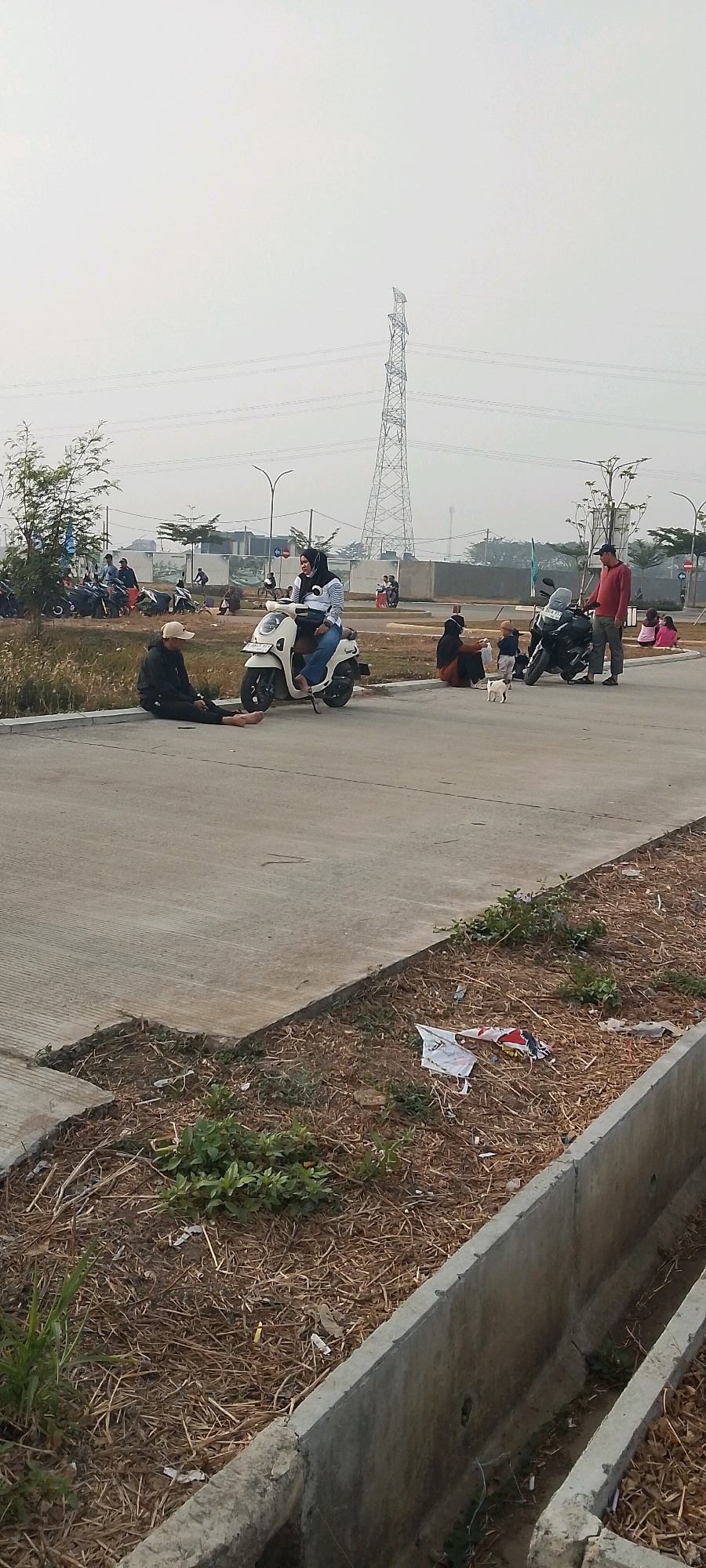 The image depicts an outdoor scene in Satriamekar, Indonesia, likely a public area with paved surfaces and undeveloped land. In the foreground, a concrete drainage ditch runs along the right side, filled with dry grass and some scattered litter. A paved path occupies the middle ground, where several people are present.
On the left side of the path, a person wearing a black hoodie and a beige cap is seated on the ground, facing away from the camera. Nearby, a white scooter is parked, and a woman wearing a hijab and striped shirt is seated on it, facing the seated person.
Further along the path, a group of individuals are gathered. One person, wearing a red shirt and a cap, stands near a black motorcycle, looking towards the right. Next to them, a woman in a hijab is seated, with a small child standing beside her. A white cat is visible in front of the child. Behind this group, another black motorcycle is parked.
In the background, there are more people scattered around, some near parked motorcycles. A tall electricity pylon with power lines is a prominent feature in the sky, indicating an urban or semi-urban setting. The sky is overcast, suggesting a hazy or cloudy day. The environment appears somewhat unkempt, with dry vegetation and visible litter.