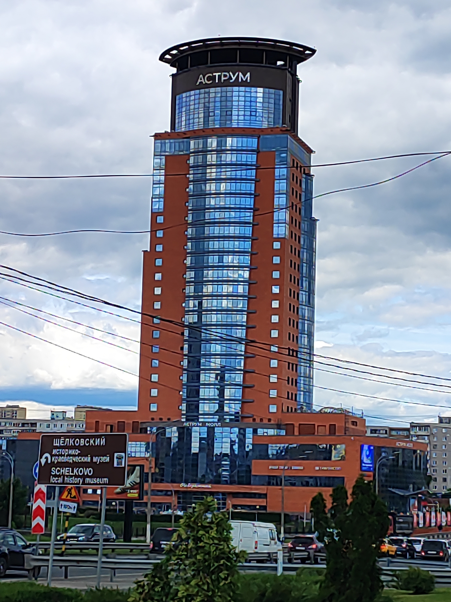 The image depicts the modern "Astrum" building in Shchyolkovo, Russia, under a sky with partially cloudy weather. The tower, featuring a combination of brown-toned exterior and reflective blue windows, rises prominently in the frame. Below the skyscraper are traffic signs, including one indicating the location of the Schelkovo Local History Museum. Below the tower is heavy traffic including a white van. In the foreground, shrubbery is present. The surrounding landscape features other urban structures, and power lines cut across the frame.