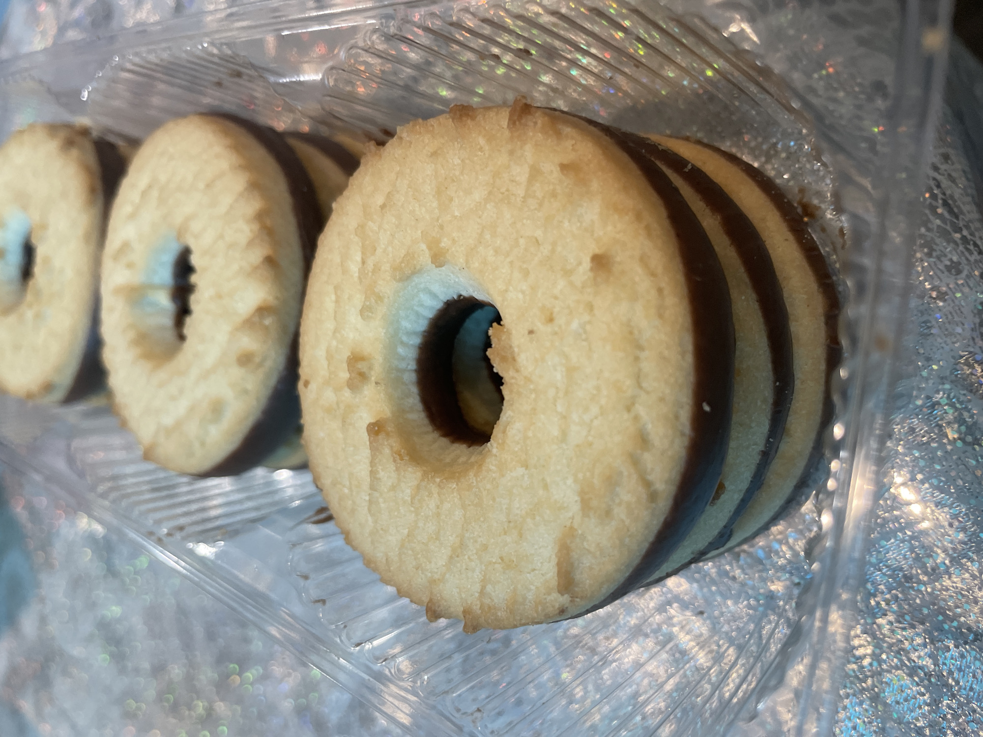 A close-up shot shows several round, cream-colored cookies with a central hole, stacked and arranged within a clear plastic container. The cookies have a distinctive dark-brown filling visible through the holes. The setting appears to be indoors, with a reflective, shimmering surface underneath the container. The location of Olginka, Russia is not explicitly apparent in the image but has been included as requested.
