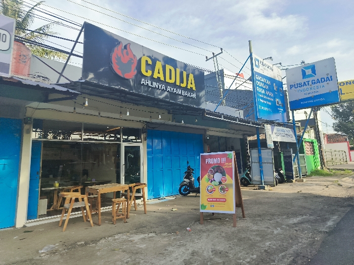 The image shows the storefront of a restaurant called "Cadja," which is located at -6.748 latitude and 107.446 longitude, in Cikalong, Indonesia. The restaurant has a blue facade and an awning with a sign that reads "Cadja Ahlinya Ayam Bakar." There is a table with three chairs outside the restaurant, and a motorcycle parked on the street. A sign for "Promo III" is visible in front of the storefront, with a menu featuring food items. There are several other signs on adjacent buildings, including one for "Pusat Gadai Indonesia." The street is paved and appears to be a commercial area. There is greenery present, including shrubs and trees.