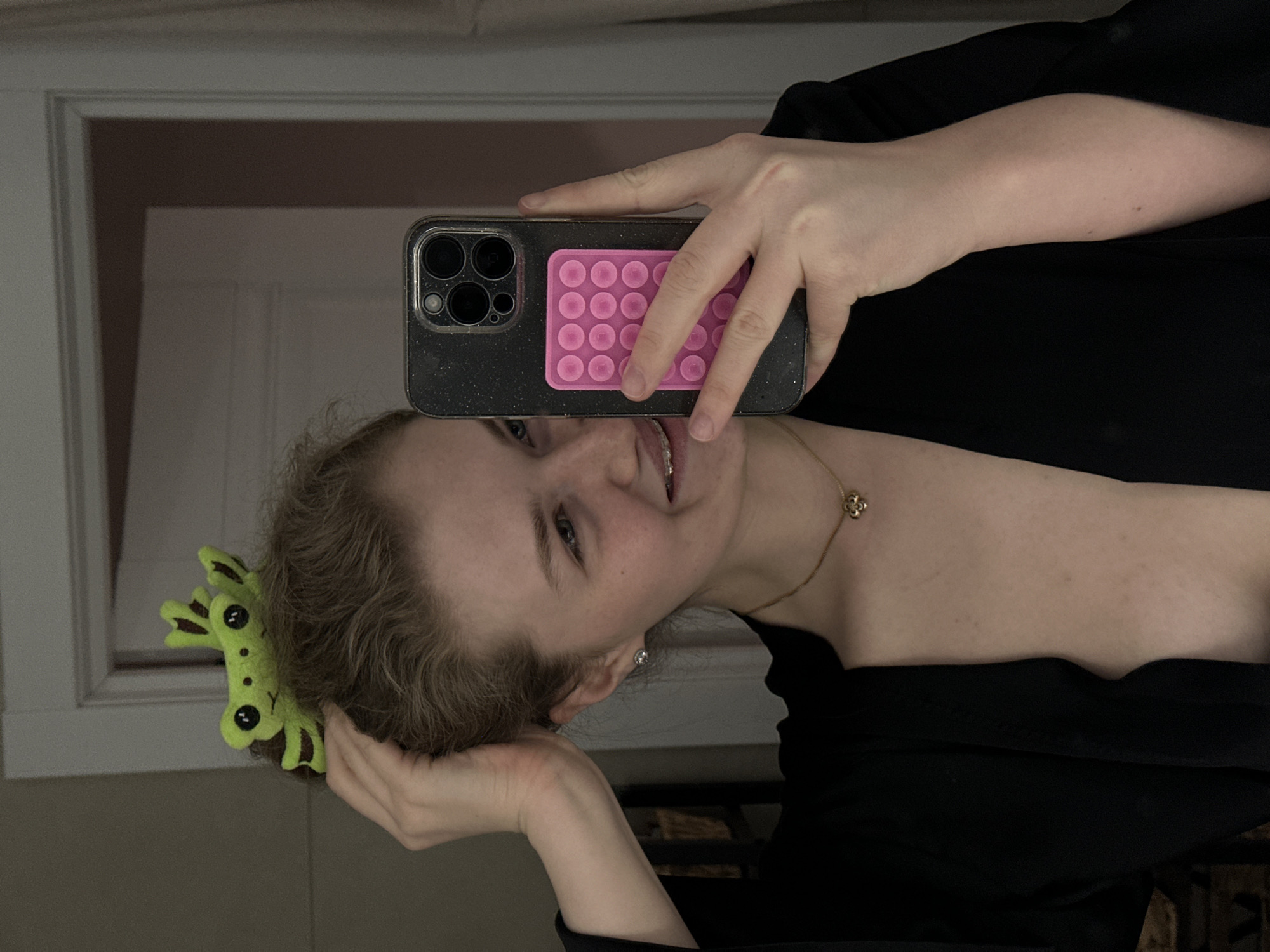 A mirror selfie shows a fair-skinned person with light brown hair, wearing a black top and a gold necklace with a flower pendant. They are holding a smartphone with a black, glittery case and a pink “pop it” fidget toy attached to the back. A green frog-shaped hair clip is visible in the person’s hair. The background includes a white door frame and a section of a wooden lattice, possibly part of a shelf or furniture. The location and city cannot be determined from this image.