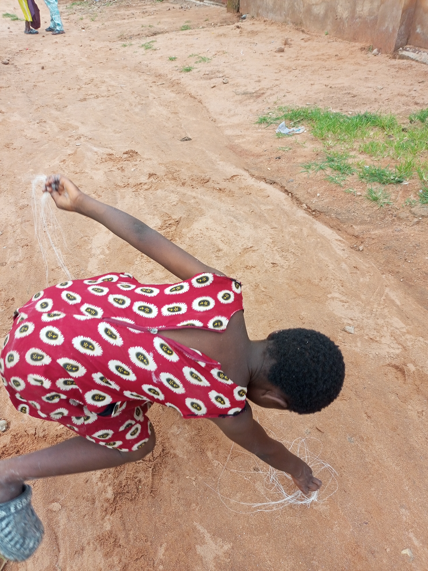 A child wearing a red and white patterned dress is kneeling on sandy ground, drawing with white string in Tempure, Nigeria. The child's hair is dark and curly, and their skin is dark. The string appears to be thin and loosely coiled on the ground. In the background, two other people are visible from the waist down, wearing light-colored pants. The ground is a mix of sand and dirt, with some patches of green grass and a partially visible concrete wall. A discarded plastic wrapper is also present on the ground.