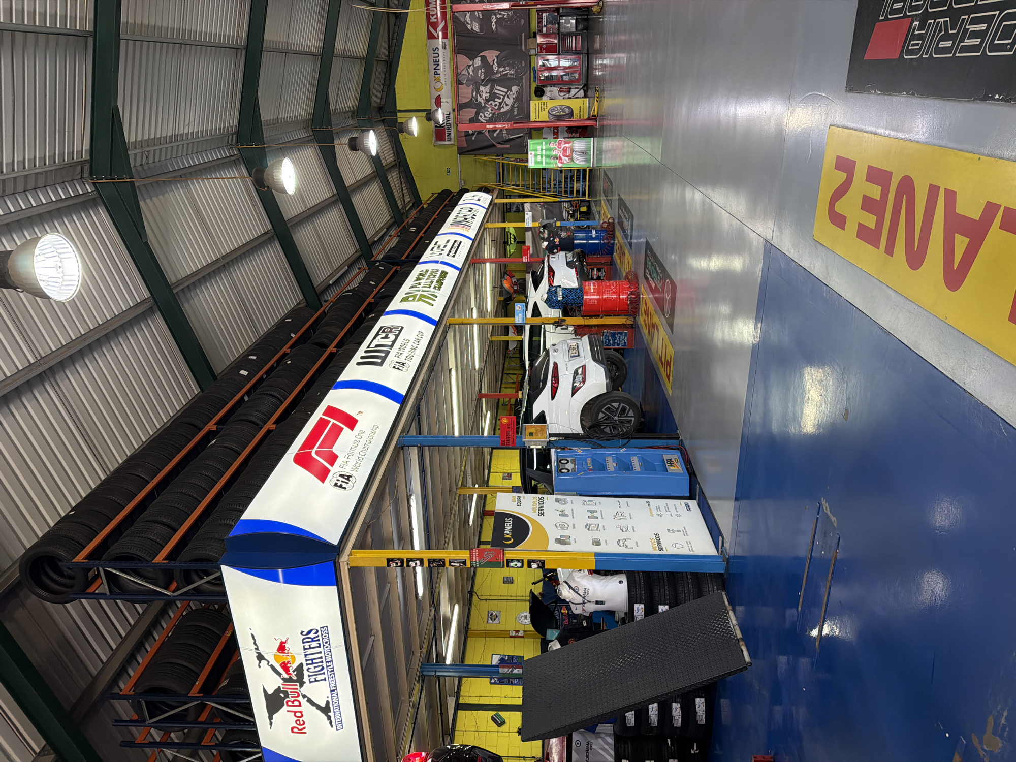 The image depicts the interior of a car service center or garage, likely in Vila Real, Portugal, given the Portuguese text on signs. The structure has a high ceiling with industrial lighting and a metal roof. Along one wall, there's a rack stacked with tires, next to a sign promoting Red Bull Fighters and FIA Formula One. Vehicles are positioned on lifts for servicing, with various tools and equipment nearby, including a blue tire inflation machine. The garage floor is a combination of gray, yellow, and blue. "LANE 2" is written on the floor. At the back of the garage, there's a promotional poster for Kumho Pneus and Red Bull.