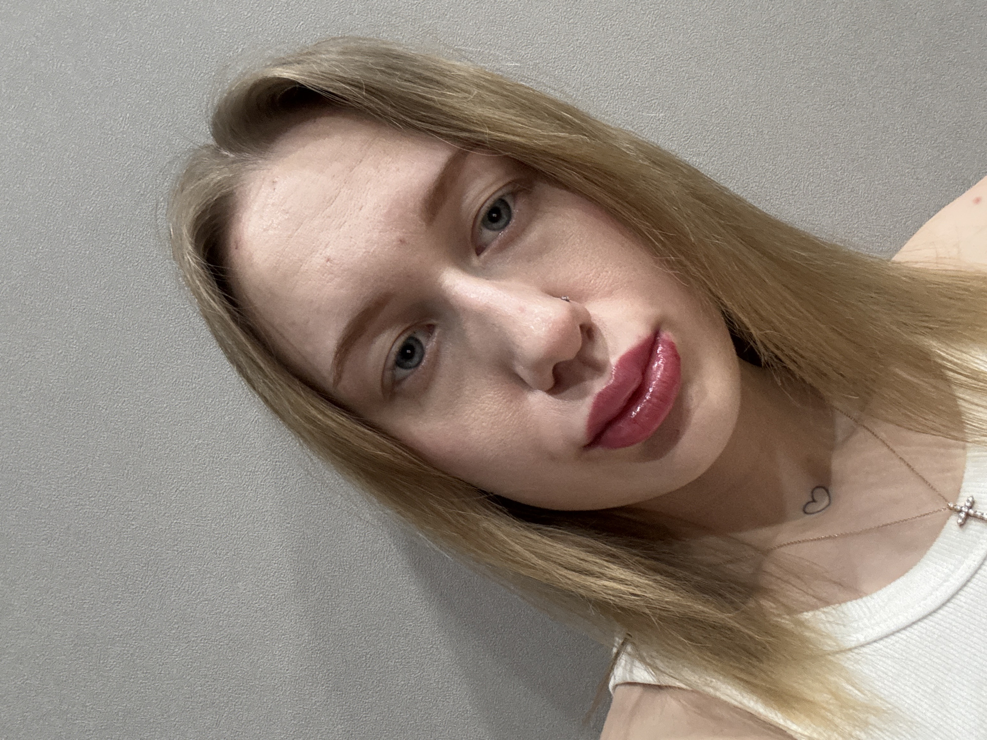 A young adult female is depicted in a close-up portrait, facing forward with her head slightly tilted to the right. She has long, straight blonde hair, parted in the middle. Her eyes are blue, and she is looking directly at the camera. A small silver stud is present in her left nostril. Her lips are full and coated with glossy, dark pink lipstick. A small, black outline heart tattoo is visible on the front of her neck, slightly to the left, below her chin. She is wearing a white ribbed tank top. A thin, gold-colored chain necklace with a small, clear gemstone-studded cross pendant is around her neck. Her skin complexion is fair, with minor imperfections visible on her forehead and left cheek. The background is a plain, light grey wall with a subtle textured finish. The precise location within Rostov-on-Don, Russia, or any specific city, cannot be determined from the visual information provided.