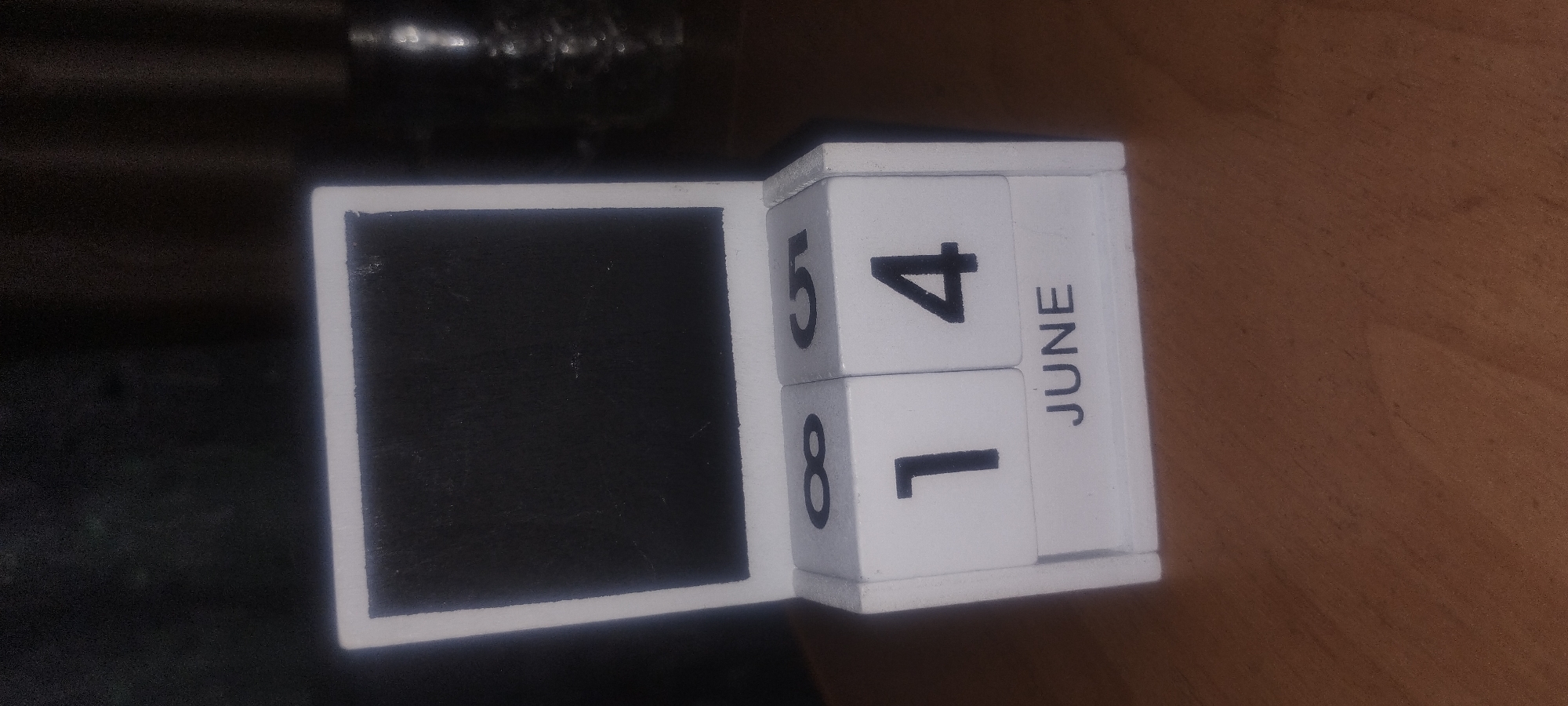 The image shows a wooden perpetual calendar with two cubes displaying the date "14" and the month "JUNE". The calendar has a black rectangular frame at the top. The calendar is placed on a wooden surface. The background is dark and out of focus, with some blurry reflections visible. There are no people or animals in the image. The image is a close-up shot of the calendar. There is no information in the image that indicates it is located in Zaporizhzhia, Ukraine.