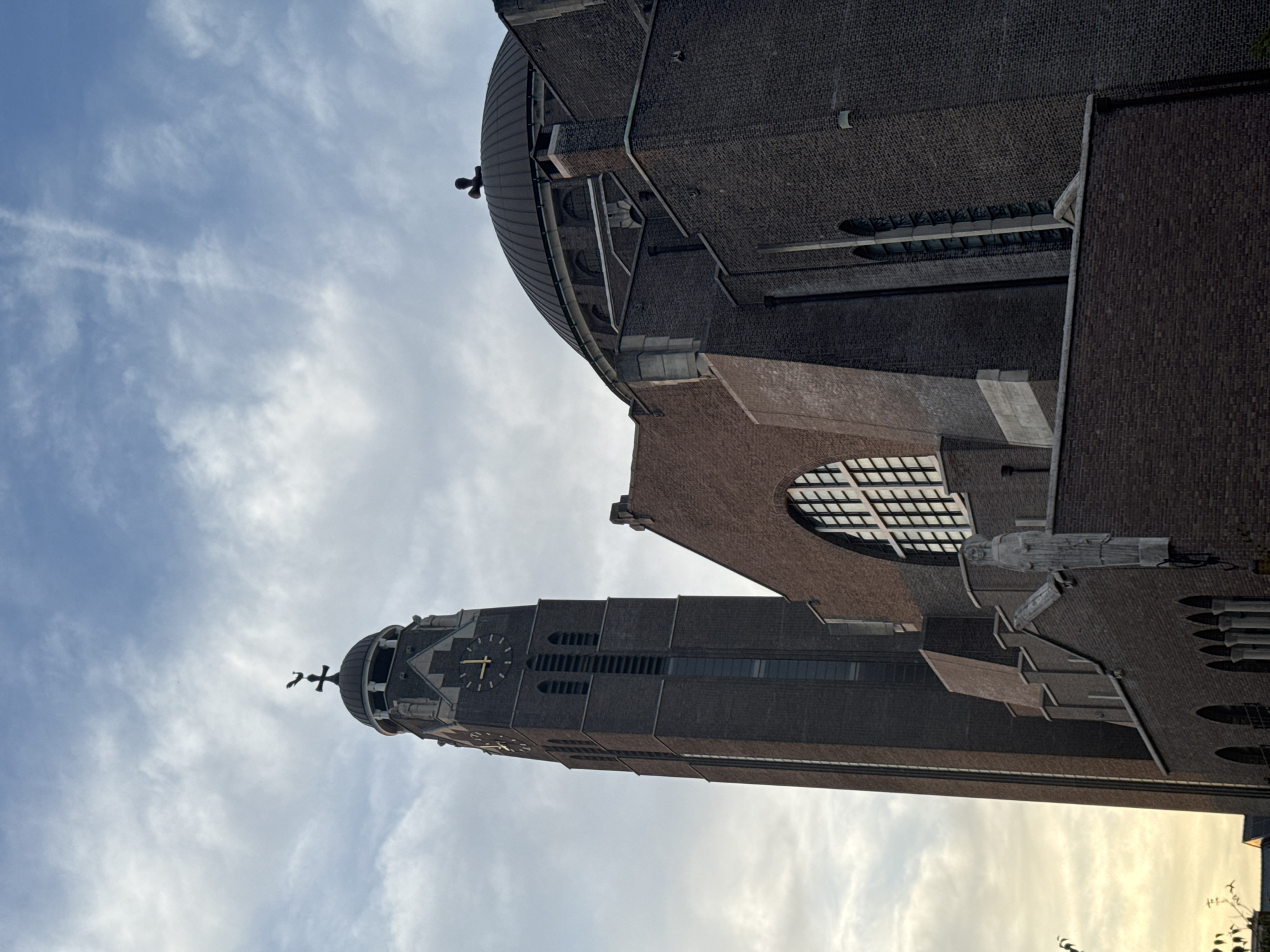 The image shows the Onze-Lieve-Vrouwekathedraal, also known as the Cathedral of Our Lady, located in Antwerp, Belgium. The viewpoint is from a low angle, looking up at the cathedral's imposing brick architecture. The sky above is partly cloudy with a soft, diffused light, suggesting it is either early morning or late afternoon.
The prominent features of the cathedral visible are its tall, dark brick bell tower on the left, topped with a cross and a weather vane. The tower has several windows and a large clock face. To the right of the tower is the main body of the cathedral, characterized by its large dome, also topped with a cross, and more brickwork. A large, arched window with intricate panes is visible on the main structure. The exterior walls are made of dark, reddish-brown brick, with some lighter stone accents, particularly around the arched window and on decorative elements of the tower. A statue of a figure is mounted on a ledge on the side of the building. The scene is devoid of people or any apparent action, focusing solely on the architectural grandeur of the cathedral against the sky. The overall impression is one of solemnity and historical significance.