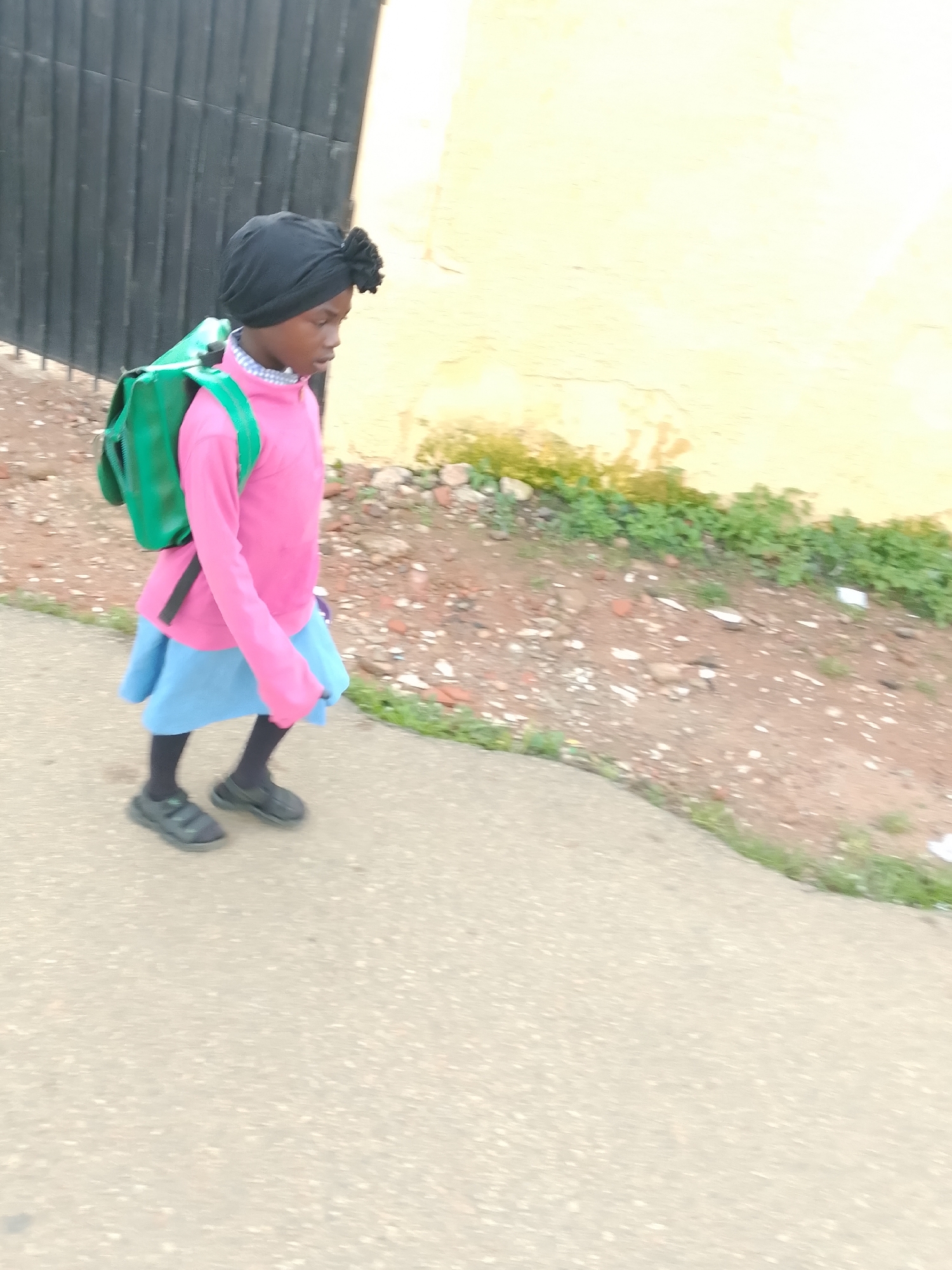 A young girl with dark skin, wearing a black headscarf and a pink long-sleeved shirt over a blue skirt, walks on a paved path. She carries a green backpack. She is wearing black socks and dark-colored shoes. To her left is a black metal fence and a light yellow wall. The ground to her right consists of dirt and sparse green vegetation. The scene appears to be in an outdoor urban environment, possibly in Jos, Nigeria, given the lighting and architectural elements.