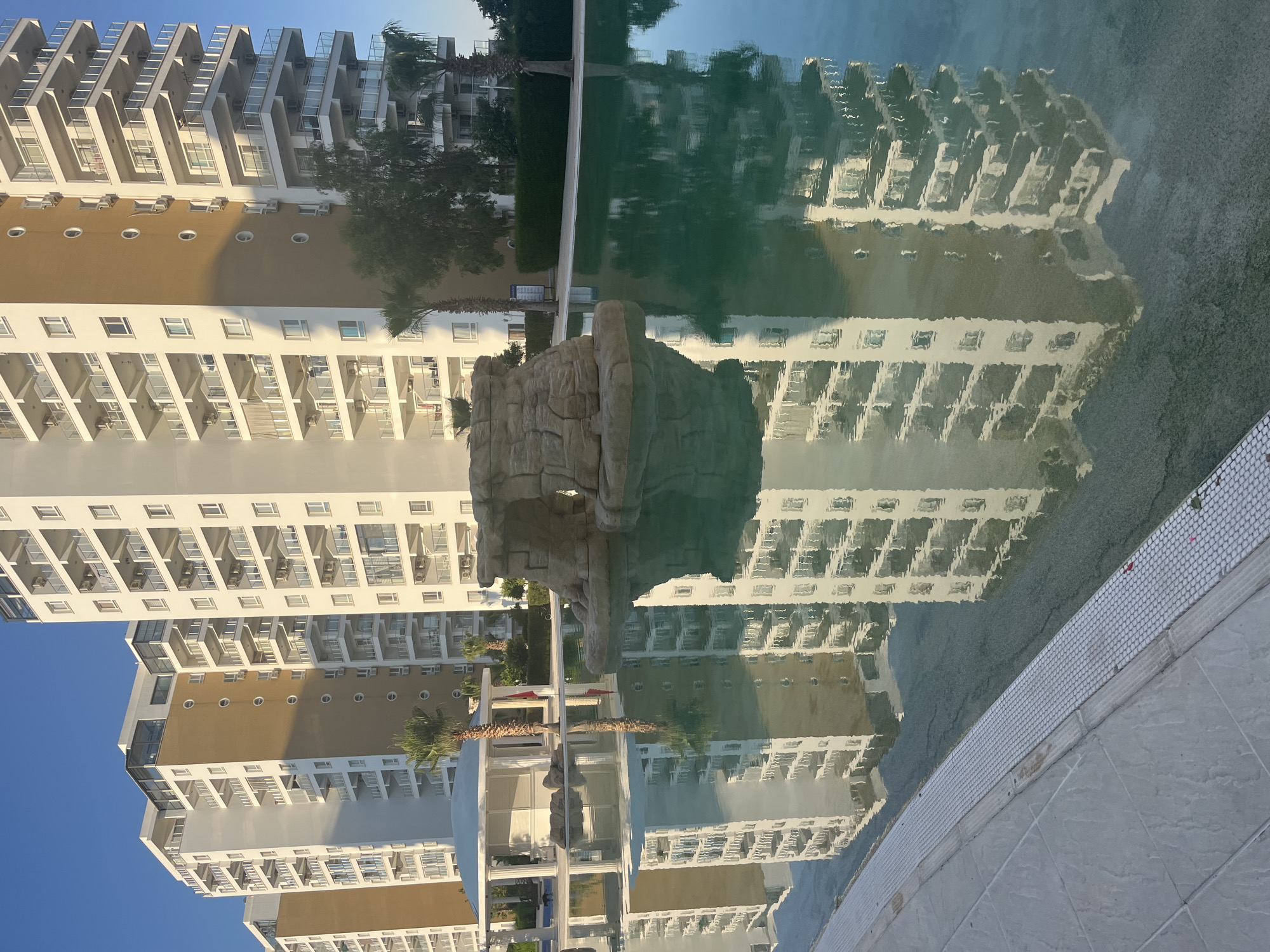 The image captures a large outdoor swimming pool reflecting several multi-story apartment buildings under a clear blue sky in Trikomo, Northern Cyprus / Cyprus. The buildings feature predominantly white facades with numerous balconies, some equipped with glass railings and visible air conditioning units. One building facade section is tan/light brown and characterized by a vertical row of small oval windows. The pool water is a teal-green color, exhibiting clear reflections of the buildings, palm trees, and the sky.
In the foreground and mid-ground, a decorative, natural-looking rock formation, likely a small grotto or fountain feature, rises from the pool. To the left, a white, circular gazebo-style structure with columns and a domed roof stands beside the pool, reflecting in the water. Several palm trees and other green foliage are visible around the pool area, some also casting reflections. The pool's edge is lined with light-colored tile work. The overall scene is devoid of people.