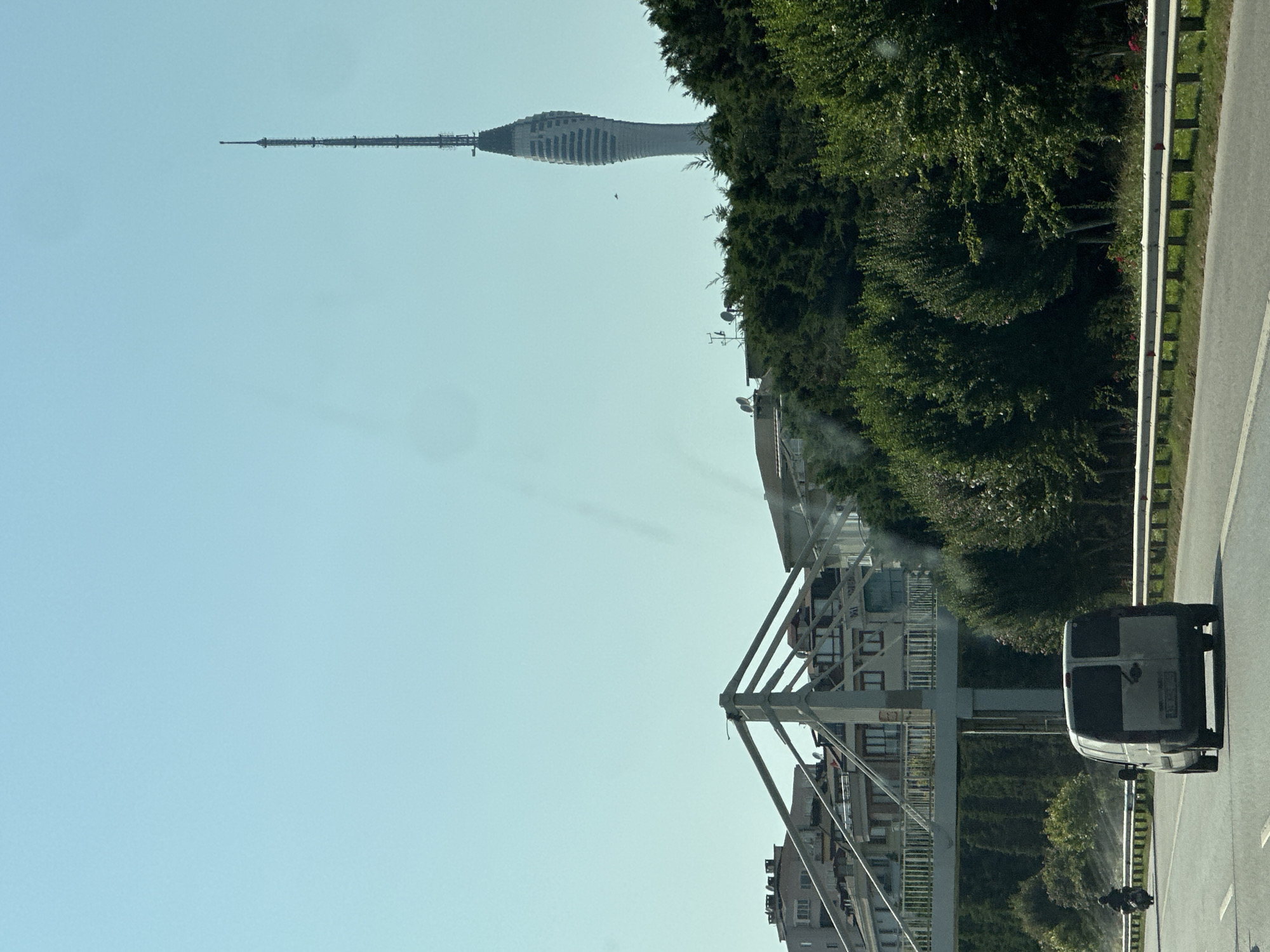 The image captures a view from a vehicle traveling on a highway in Ataşehir, Turkey. A white van is in the foreground, driving on the right side of the road. To the left of the van, a person on a motorcycle is also on the highway.
In the middle ground, a pedestrian bridge spans across the highway. Behind the bridge, buildings with multiple floors are visible. A tall, slender tower with a distinct curved design and antenna at the top dominates the background, rising above a dense line of green trees. The sky is clear and blue. The scene depicts a typical urban highway environment with infrastructure and residential buildings.