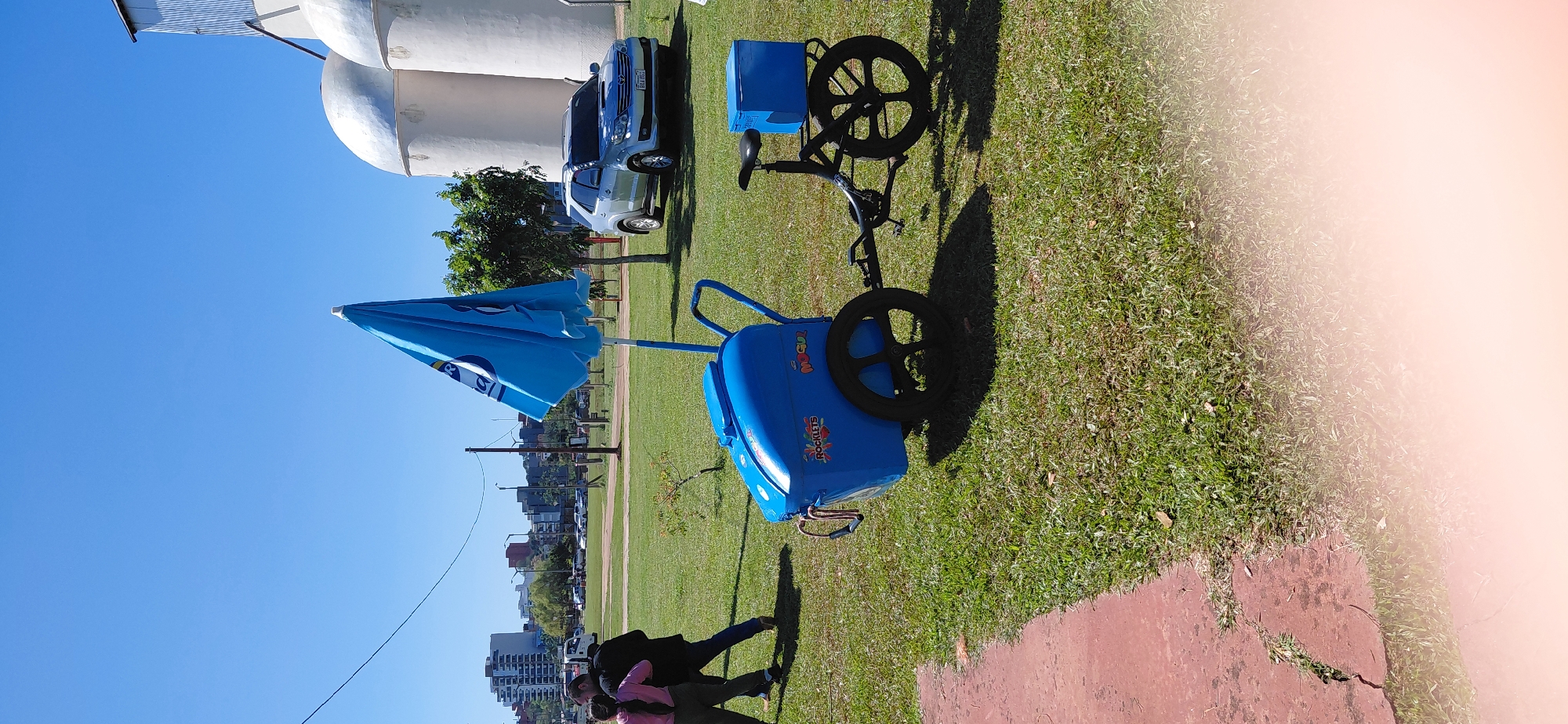 In Encarnacion, Paraguay, on a sunny day, a blue ice cream cart with a closed blue umbrella is parked on a grassy area. The cart has "ROCKLETS" and "MOGUL" written on its side. Attached to the cart is a bicycle with a blue cooler box on the back. In the background, a silver SUV is parked, and beyond that, a cityscape with buildings is visible. Two people, one in a pink shirt and dark pants, and another in a dark jacket and pants, are walking away from the viewer on the grass. The environment is a park-like setting with green grass and some trees.