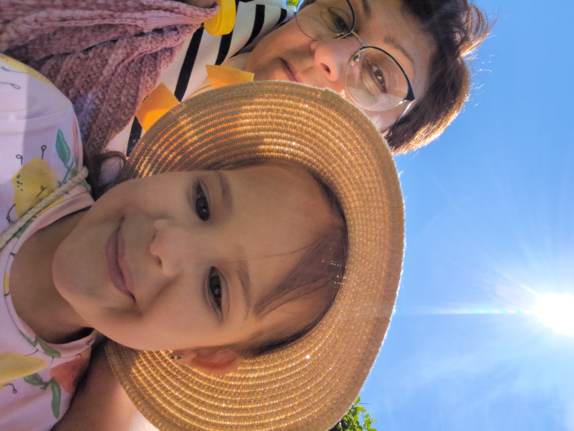 This image captures a bright, low-angle selfie featuring an adult woman and a young child, likely outdoors in Sochi, Russia, under a clear blue sky. The perspective is from below, looking up at the subjects.
In the upper part of the frame, an adult woman with short, dark hair and glasses looks towards the camera with a calm expression. She is wearing a black and white striped top layered with a textured pink or purple garment.
Dominating the lower portion of the image is a young child, positioned closer to the camera. The child is wearing a straw hat that covers most of their forehead but reveals a sweet smile and bright eyes. Their top is light-colored with a colorful fruit pattern, possibly lemons or oranges.
The background is a vibrant, cloudless blue sky, indicating excellent weather. A strong sun flare is visible in the upper right corner, suggesting bright sunshine, likely mid-day or afternoon. A hint of green foliage is present in the lower right, further confirming an outdoor setting. The overall mood is cheerful and relaxed, reflecting a pleasant day spent together. No text is visible in the image.