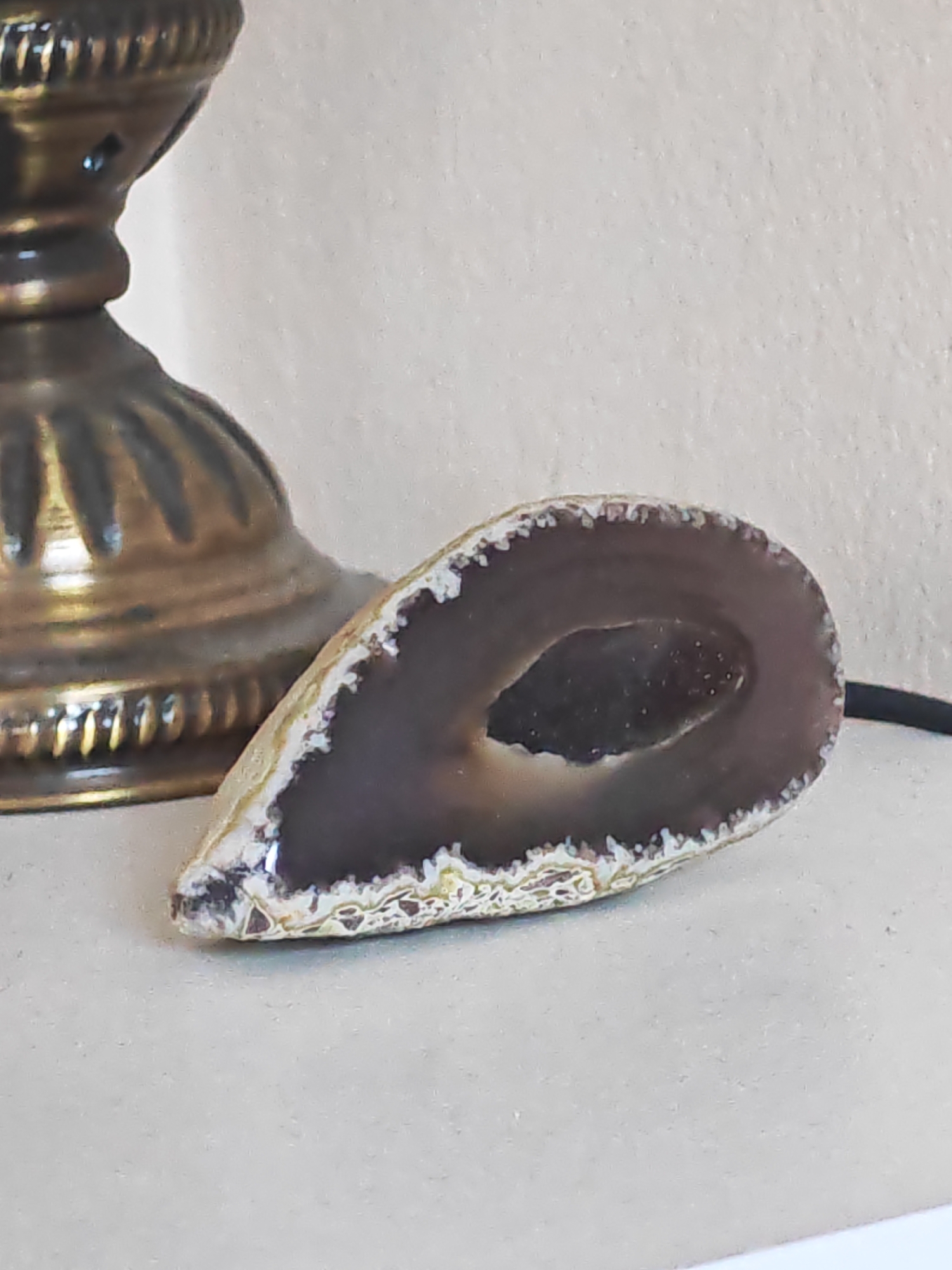 A close-up shot shows a sliced geode with a dark purple interior and a white, crystalline border. The geode rests on a light-colored surface. To the left of the geode, partially visible, is the base of a brass lamp with intricate detailing. The background is a textured, off-white wall. The image appears to be taken indoors.