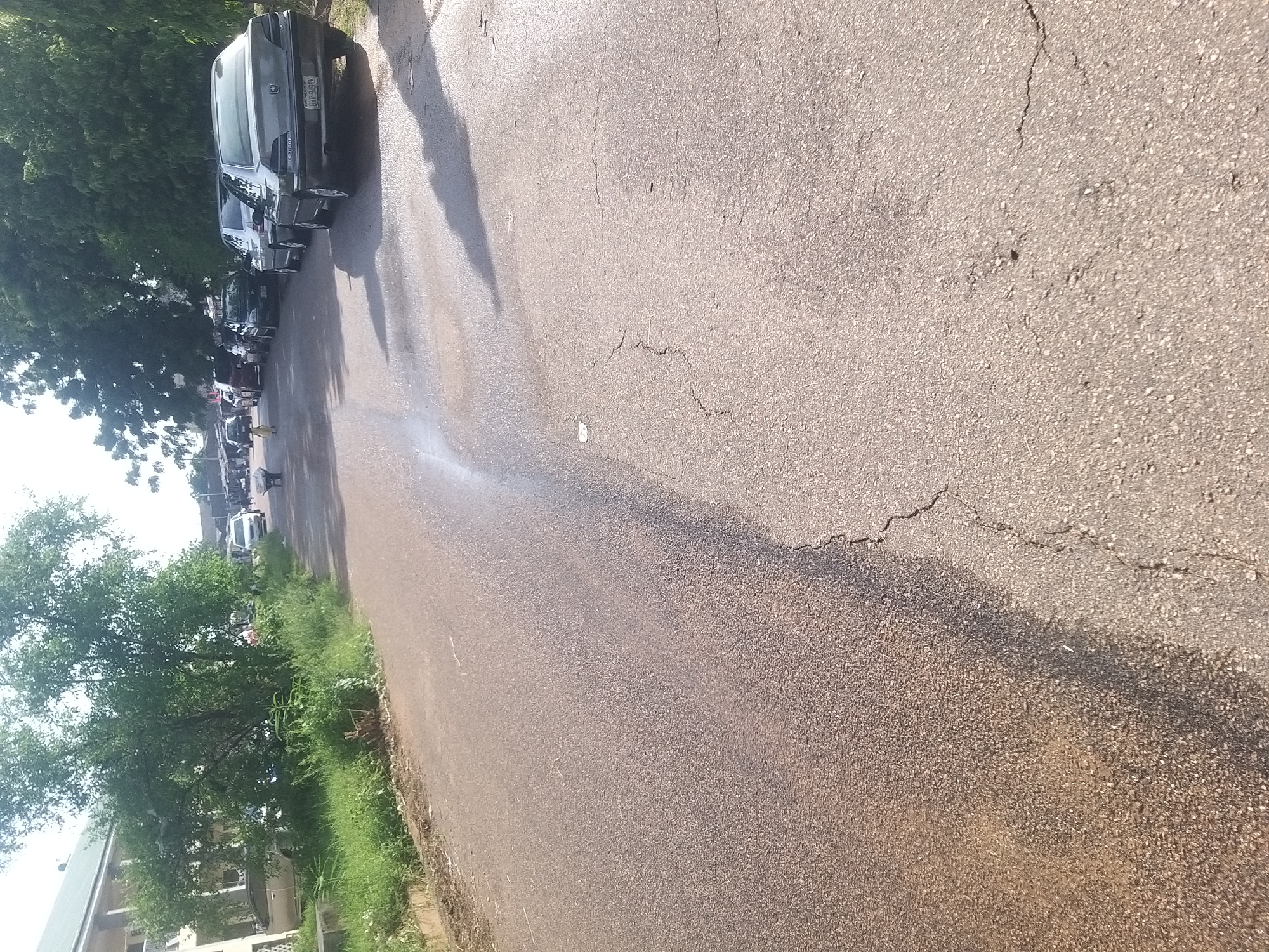 The image shows a street scene in Kano, Nigeria. A grey Peugeot sedan is parked on the right side of the road, with its license plate visible as BXC-949KK. Several other cars are parked along the street, and some trees line the side of the road. A person in a yellow dress is walking on the left side of the street. In the background, a white van and another vehicle are visible. The road appears to be paved with asphalt and shows signs of wear, with cracks present. The overall environment suggests a typical urban street in Nigeria.