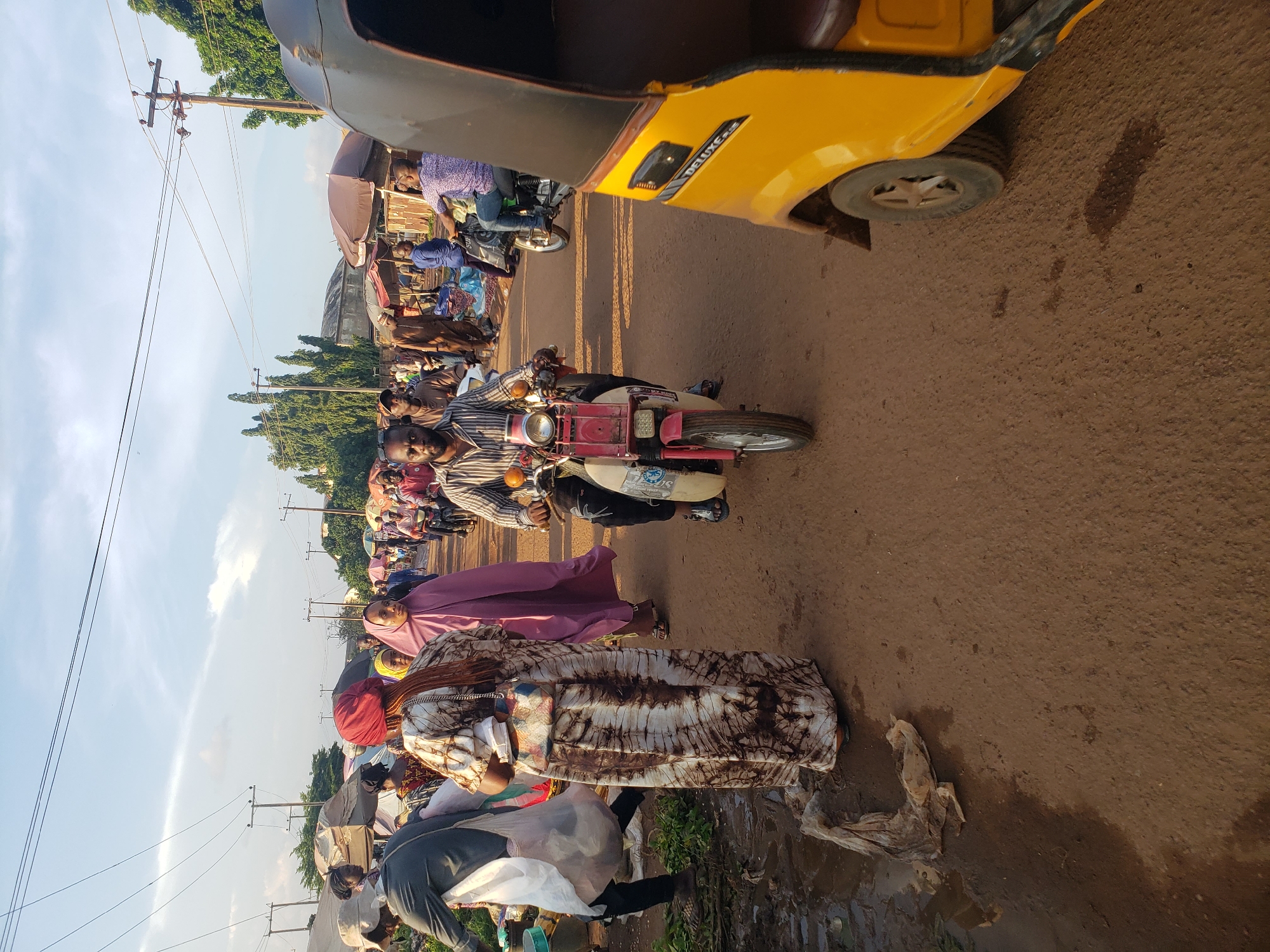 In the city of Kakuri Gwari, Nigeria, a bustling street market scene unfolds. The ground is a mixture of reddish-brown earth and puddles, indicative of recent rain or water spillage. A yellow auto-rickshaw, possibly a commercial taxi, is partially visible in the foreground on the right, its front wheel and part of its body painted yellow with black trim. Further down the dusty road, a man rides a red and white scooter, wearing a striped shirt and dark pants. Behind him, another man on a motorcycle, wearing a patterned blue shirt and jeans, is also present. The market is populated by various individuals. To the left, a person in a grey jacket carries a white plastic bag. A woman in a distinctive tie-dye patterned dress stands in the mid-ground, holding a white object, and to her right, another woman wears a purple headscarf and dress. Numerous market stalls with umbrellas are visible in the background, suggesting a vibrant commercial area. Overhead, power lines crisscross the sky, supported by utility poles. The overall atmosphere is one of activity and commerce on a bright, possibly late afternoon, day.