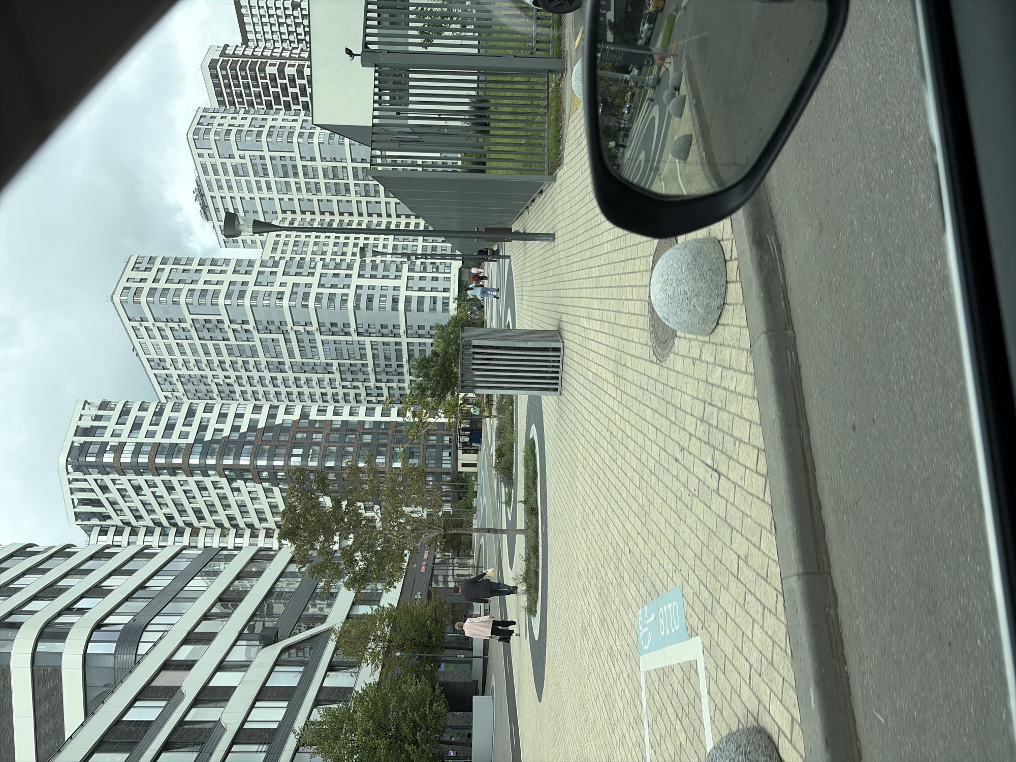 A view from a vehicle looking out at a modern residential complex in Moscow, Russia. The scene depicts tall, multi-story buildings with a grid-like facade, featuring numerous windows and balconies. In the foreground, a paved walkway with a yellow brick pattern is visible, bordered by a curb and street. Several spherical concrete bollards are placed along the edge of the walkway, and a blue handicap parking symbol is painted on the ground. A tall metal fence encloses a grassy area to the right. Two individuals, a woman in a pink coat and a man in a dark jacket, are walking away from the camera on the walkway. A street lamp stands on the walkway between them and the buildings. The sky is overcast.