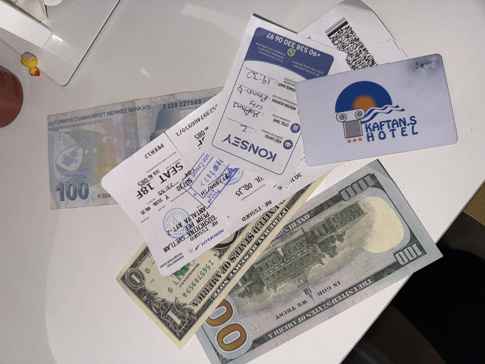 The image shows a close-up view of a white table surface with several objects scattered on it. In the foreground, a US one hundred dollar bill and a US one dollar bill are laid out. Overlapping these bills are two boarding passes, one from "AEROFOLOT" with flight details including "SEAT 18F" and "UR. 00:35", and another from "KONSEY" with handwritten notes indicating "Rotlans", "city", "Perm", and a time of "19:30". A white hotel key card for "KAFTAN'S HOTEL" is also visible. In the background, a Turkish 100 Lira banknote is partially visible, along with a small yellow duck figurine. The lighting suggests an indoor setting, possibly a hotel room or a desk. There is no explicit indication of the city of Perm, Russia, within the visible elements of the image, although one of the boarding passes has a handwritten notation that includes "Perm".