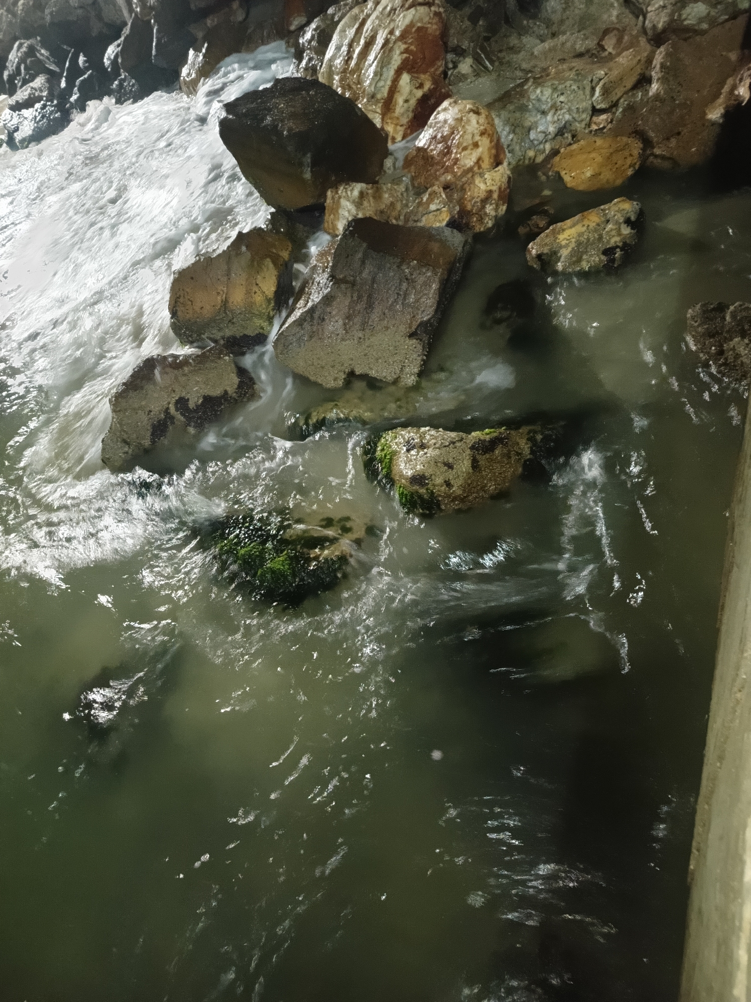 The image shows water flowing around and over rocks. The water appears to be greenish-gray and possibly turbulent. Some of the rocks are covered in moss or algae. The light suggests it is either nighttime or a very dimly lit area. The location is near a structure (possibly a wall) on the right edge of the frame. The location, Juan Griego, Venezuela, is provided, though not discernible from the image alone. No people or interactions are depicted.