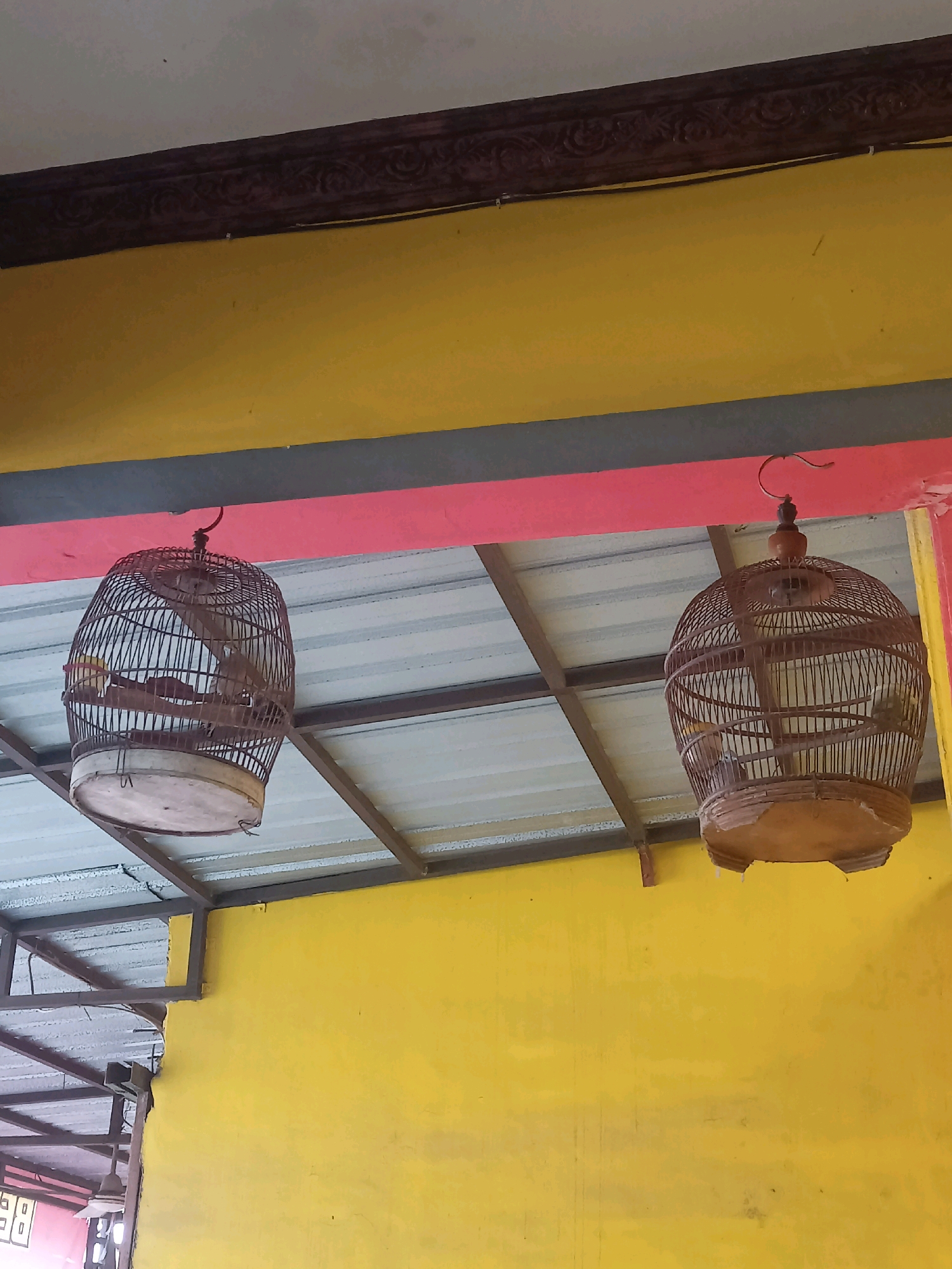 Two traditional woven bird cages are suspended from a ceiling. The cages are made of bamboo and have circular bases and domed tops with a perch inside each. They hang from hooks attached to what appears to be a metal structure supporting a corrugated metal roof. The background consists of a yellow wall beneath a dark brown decorative trim and a pink horizontal band. The scene is outdoors, likely in a sheltered area such as a porch or under an awning, indicated by the visible ceiling structure and wall. The lighting suggests it is daytime. No people are present in the image.