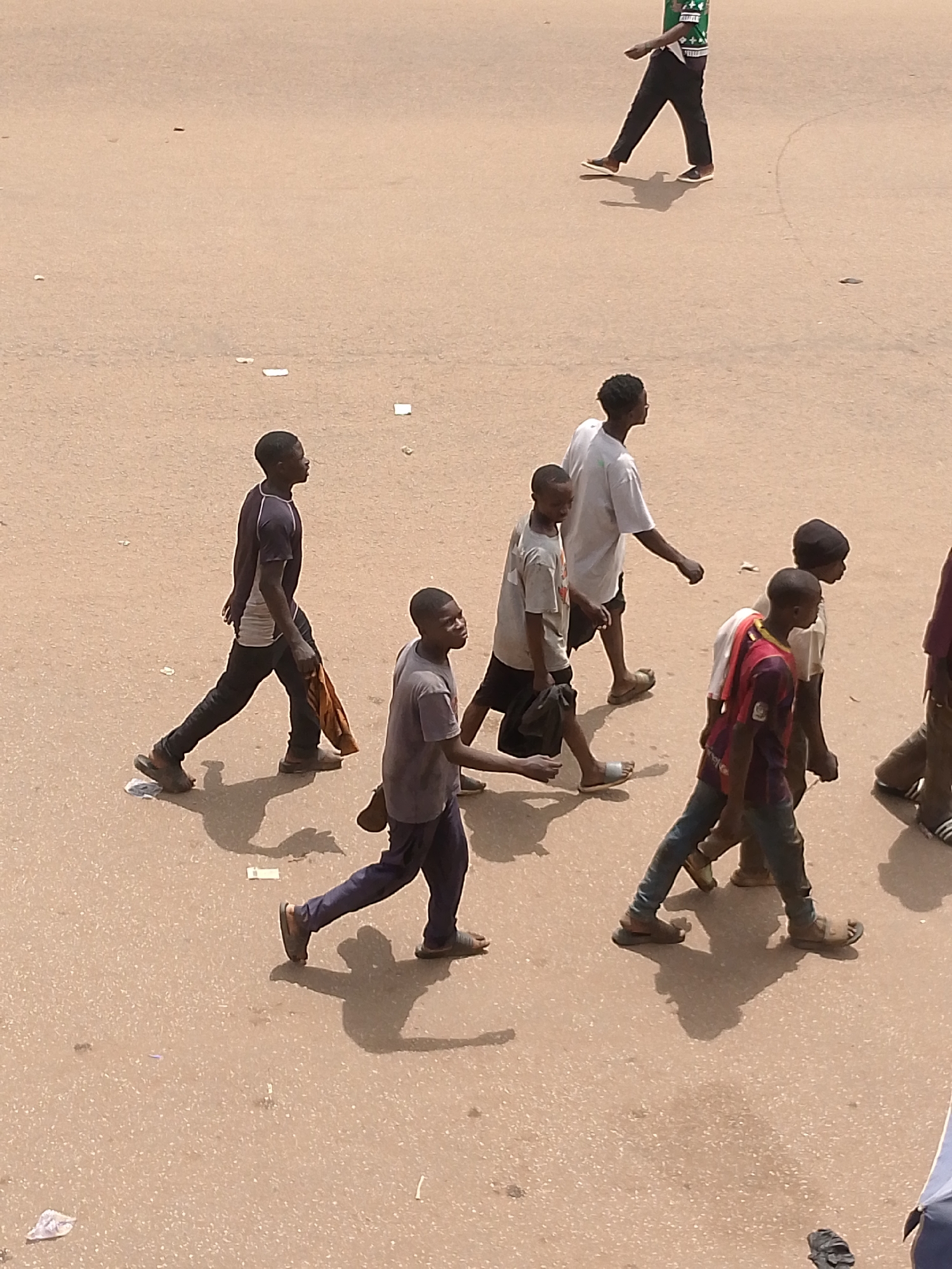 A group of young people is walking on a tan-colored street in Bukuru, Nigeria. One person is walking in the upper right corner. The rest of the group includes six people of color, dressed in various casual clothes. The group appears to be walking in the same direction. The road surface is smooth and shows some small scattered debris. The scene appears to be in daylight.