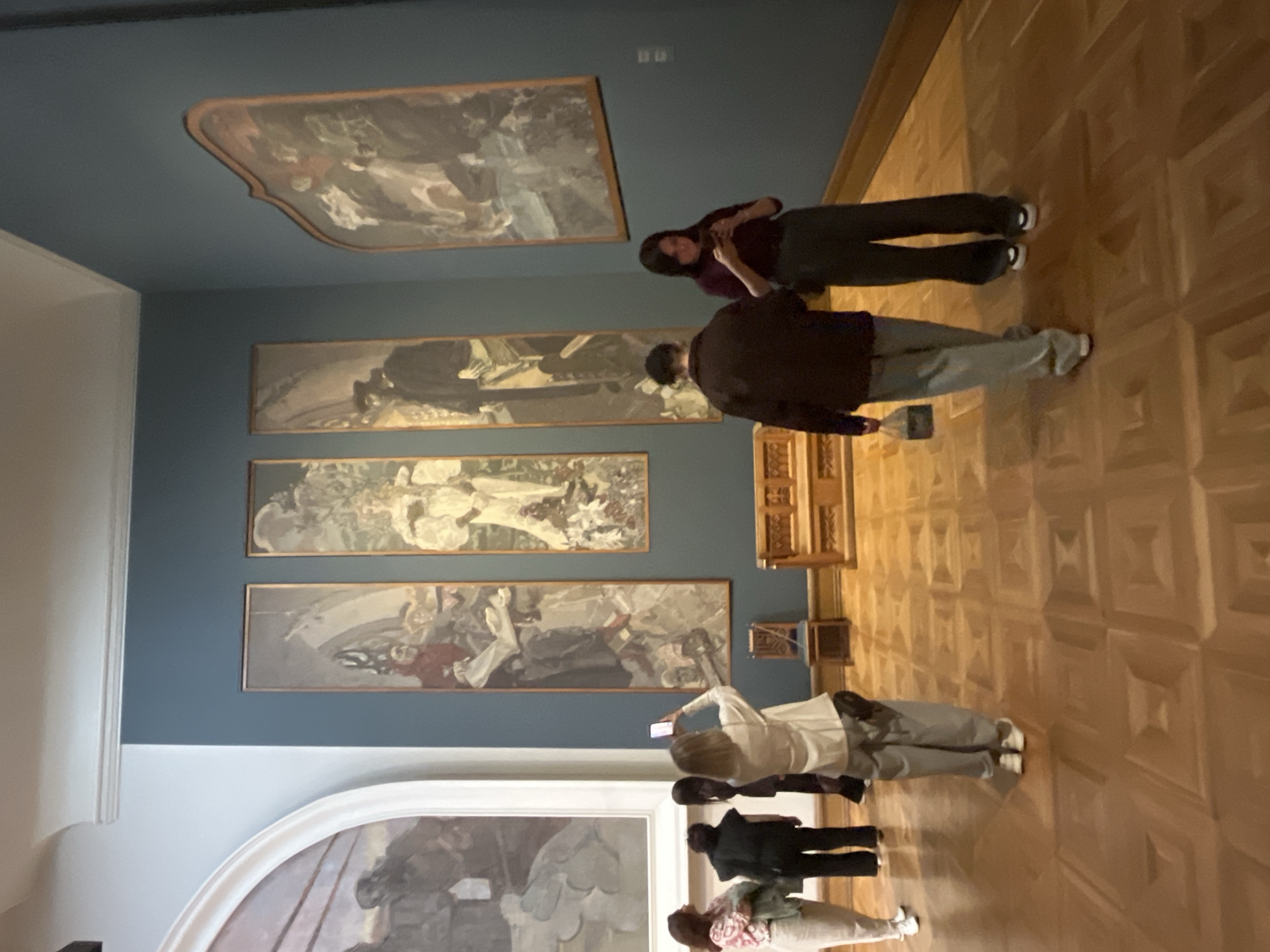 The image depicts the interior of a museum, likely the Tretyakov Gallery in Moscow, Russia, given the style of the paintings and the architectural elements. Several people are present, observing artworks displayed on a dark blue wall.
In the foreground, a person is kneeling and taking a photo with their smartphone of a tall, slender painting. To their left, three other individuals are standing, looking at a large, arched painting partially visible on the left side of the frame.
Further into the room, two more people are walking away from the camera. One is wearing a dark jacket and light-colored pants, carrying a small black bag. The other, a woman, is wearing a burgundy top and dark pants.
Several artworks are visible on the wall. Three tall, narrow paintings hang vertically, each depicting figures in historical or allegorical dress. A larger, irregularly shaped painting is mounted on the right wall. In the middle ground, a wooden bench is placed in front of the artworks, and a wooden chair is positioned near the central paintings. The floor is made of polished hardwood, arranged in a geometric parquet pattern. The lighting appears to be ambient museum lighting.