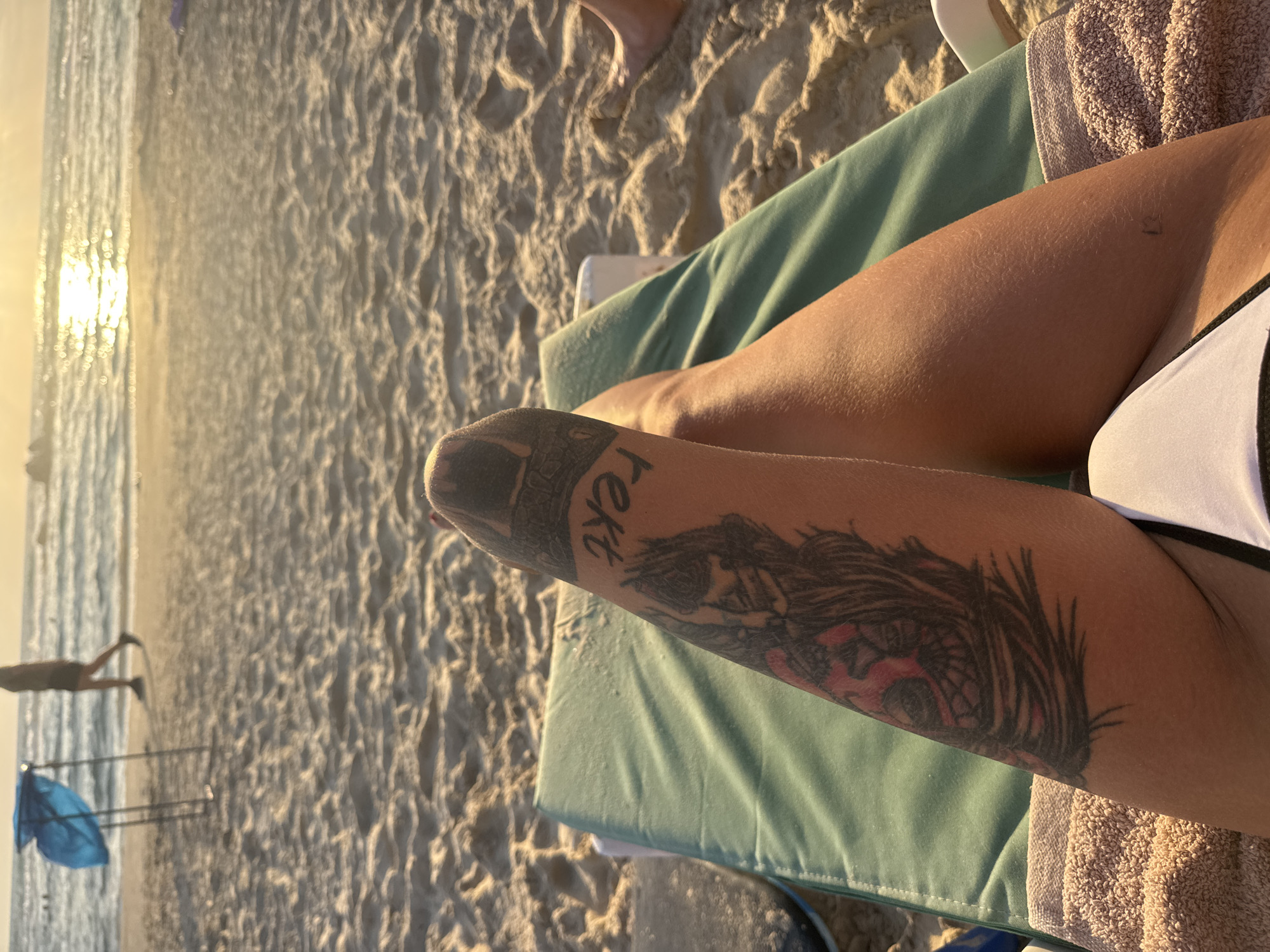 A close-up, vertical shot shows a person's bare leg with a tattoo on their forearm. The tattoo is of a face with prominent eyes and what appears to be hair or a mane. The word "Text" is visible in black ink on the upper part of the forearm, above the tattoo. The person is lying on a beach chair, which has a teal cushion and a tan towel. The background consists of sand with ripples, suggesting a beach setting. In the distance, the ocean is visible, with the sun reflecting on the water, indicating it is either sunrise or sunset. The lighting is warm and golden. Part of another person's legs and feet are visible in the top left corner of the frame, and a blue umbrella is visible in the far distance.