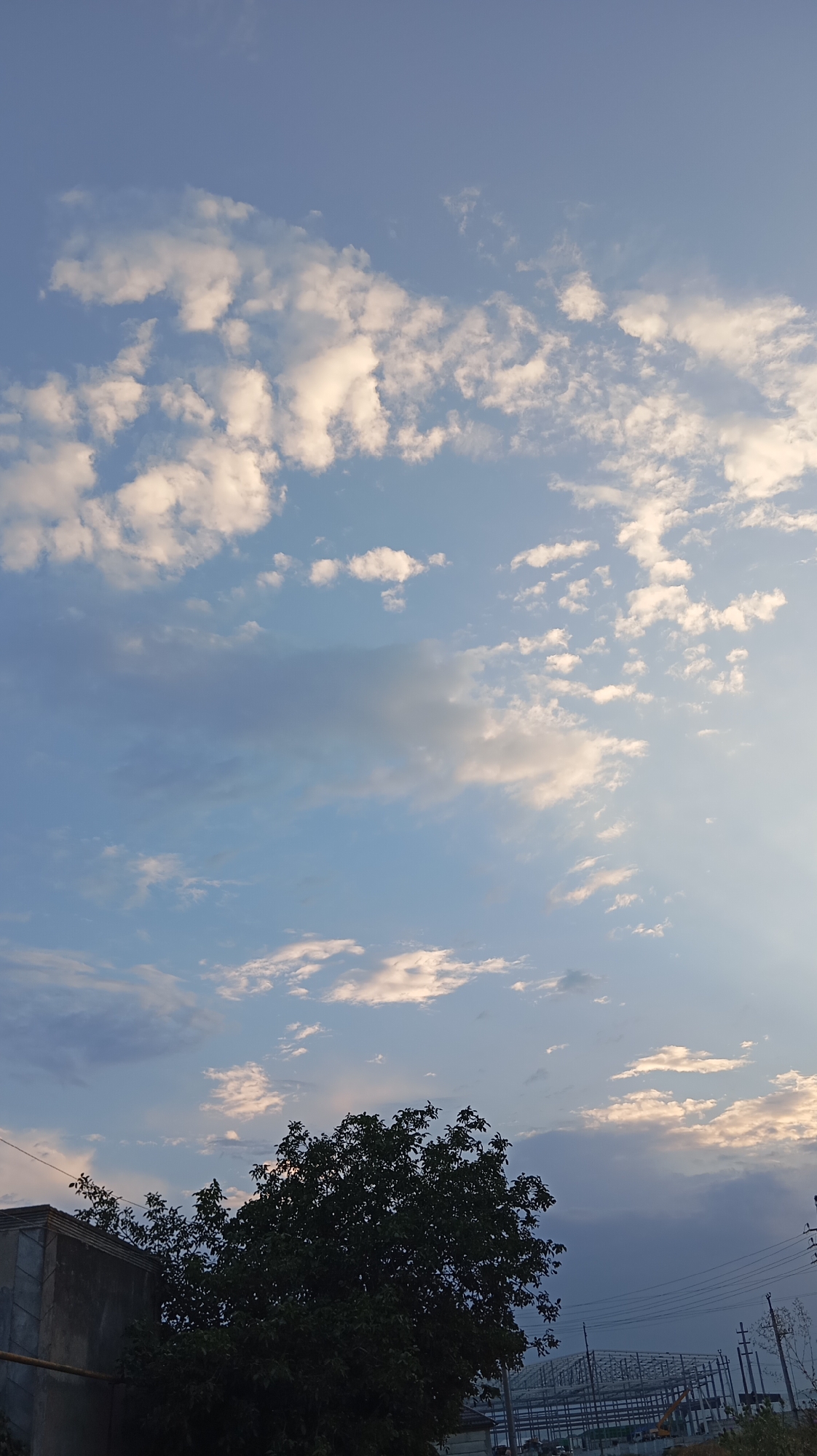 A vertical, eye-level shot captures a sky with scattered white and grey clouds, backlit by a subtle light source on the right. The lower portion of the image is dominated by the silhouette of a large, leafy tree on the left, partially obscuring a grey concrete structure with a rough texture. Beyond the tree and structure, a construction site with a steel frame building is visible, along with power lines. The scene is set in the city of Baksan, Russia.