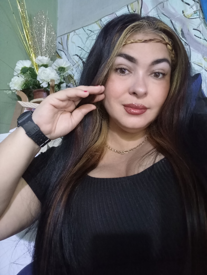 A woman with long, dark hair and a braid-like headband poses for a selfie. She wears a black, fitted top and a gold chain necklace. A large black watch is visible on her left wrist. Her makeup includes glossy lipstick. Behind her, a decorative arrangement of white flowers and gold tinsel sits on a small table. A patterned fabric or curtain serves as a backdrop.