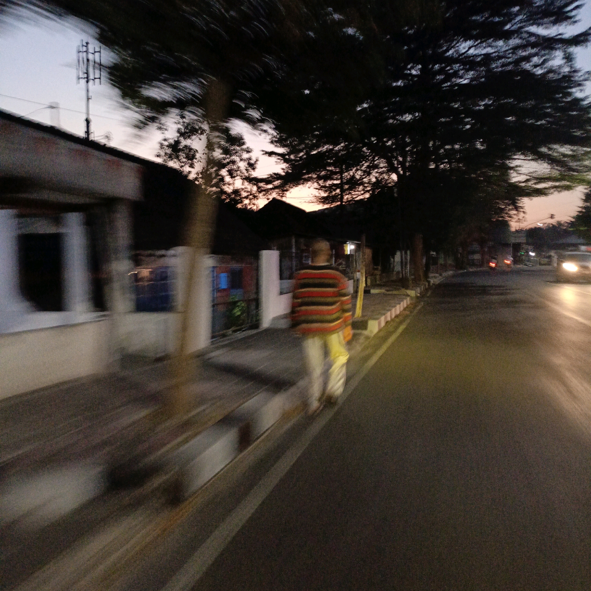 This image depicts a street scene in Salatiga, Indonesia, likely during the twilight hours due to the ambient light conditions. In the foreground, a person wearing a horizontally striped sweater in red, orange, and black, along with light-colored pants, is walking away from the camera on a sidewalk. They are carrying a yellow bag. The sidewalk is bordered by a road on the right, which appears wet and reflects light from vehicles. Buildings line the left side of the street, characterized by a dark roofline and what appear to be windows or openings. Trees with dark foliage are visible throughout the scene, some casting shadows on the sidewalk. A communication tower with multiple antennas is present in the upper left portion of the image. The overall impression is one of movement and motion blur, suggesting the photograph was taken from a moving vehicle. The sky shows a gradient of light colors, indicative of either dawn or dusk.