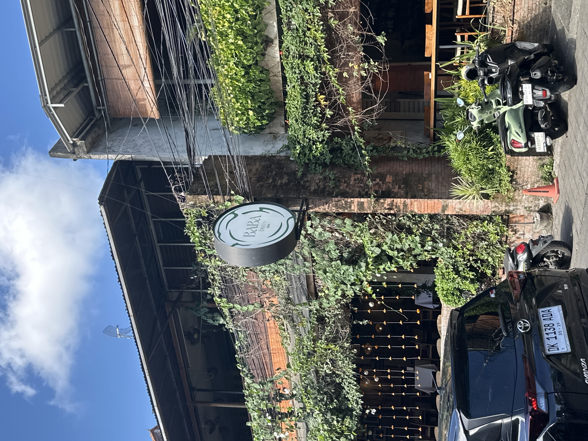 A sunny outdoor scene in Ubud District, Indonesia features a two-story building with a covered outdoor dining area on the first floor and an open-air seating area on the second. The building is covered in abundant green foliage, with vines and plants cascading down the brick walls and over the railings. A circular sign for "BABA Bistro" is prominently displayed on the side of the building.
In the foreground, a black Toyota Avanza is parked on the street, its license plate reading "DK 1138 ADA". Next to it, two scooters are parked, a light green one and a black one. The scooters have helmets on their handlebars. An orange traffic cone is positioned on the sidewalk. The street is paved with interlocking blocks, and the overall impression is one of a vibrant, tropical urban environment. The sky is blue with scattered white clouds.