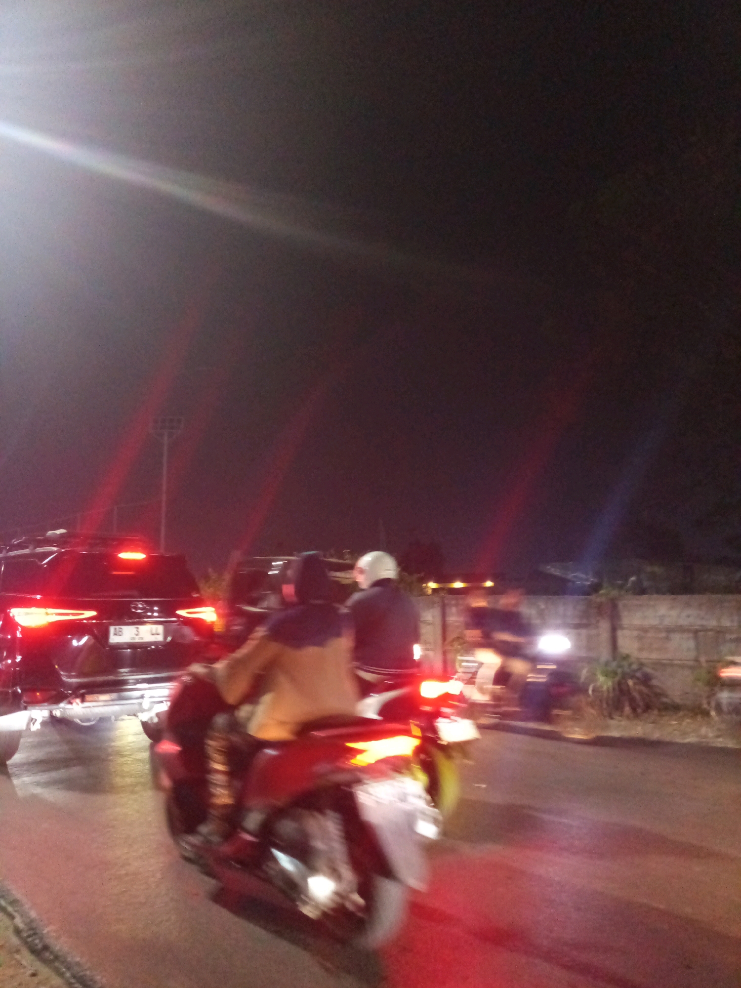 The image captures a nighttime scene of traffic congestion on a road in Bekasi, Indonesia. In the foreground, a motorcycle with two occupants is moving, with the rear rider in focus wearing a brown jacket and a dark head covering. The motorcycle's brake light is illuminated. To the left, a black SUV is stopped, its brake lights and taillights are brightly lit. The license plate of the SUV reads "AB 3 LL". Other vehicles and motorcyclists are visible further down the road, indicating heavy traffic. The environment is dark, illuminated by artificial lights from the vehicles and streetlights, creating lens flares and reflections on the wet road surface. The background shows a low wall and some vegetation.