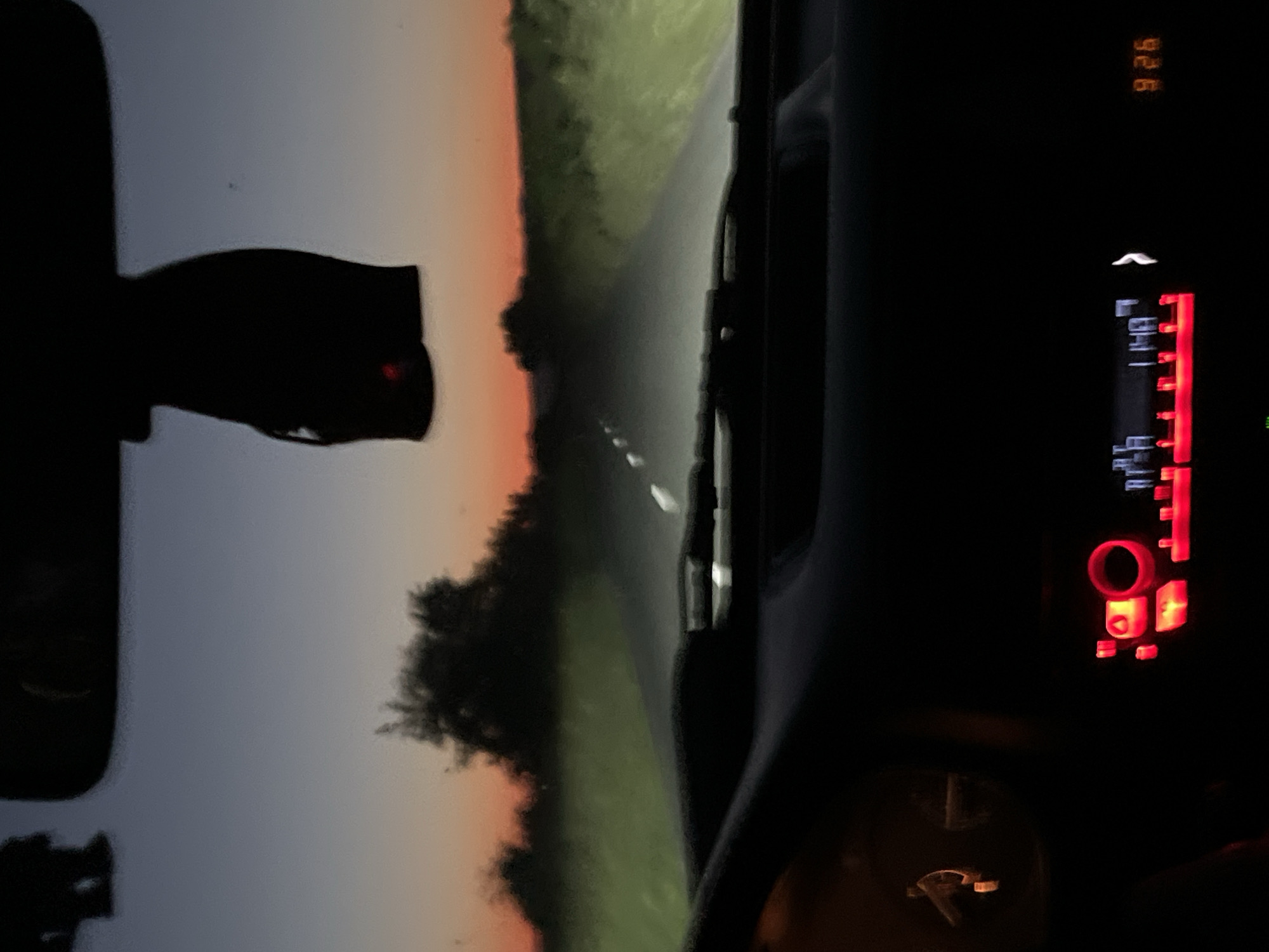 The image shows a view from inside a vehicle, likely a car, driving on a road at dusk or dawn. The road appears to be rural, bordered by grassy areas and trees. The sky is transitioning from daylight to darkness, indicated by the gradient of orange and grey. The rearview mirror is visible on the left, reflecting a blurry image. The dashboard of the vehicle is visible on the right, showing the car radio with red illuminated buttons and displays. A speedometer is also visible on the dashboard. The time displayed on the radio is 9:26. The location of Tankovo, Bulgaria, is not discernible from the visual information.