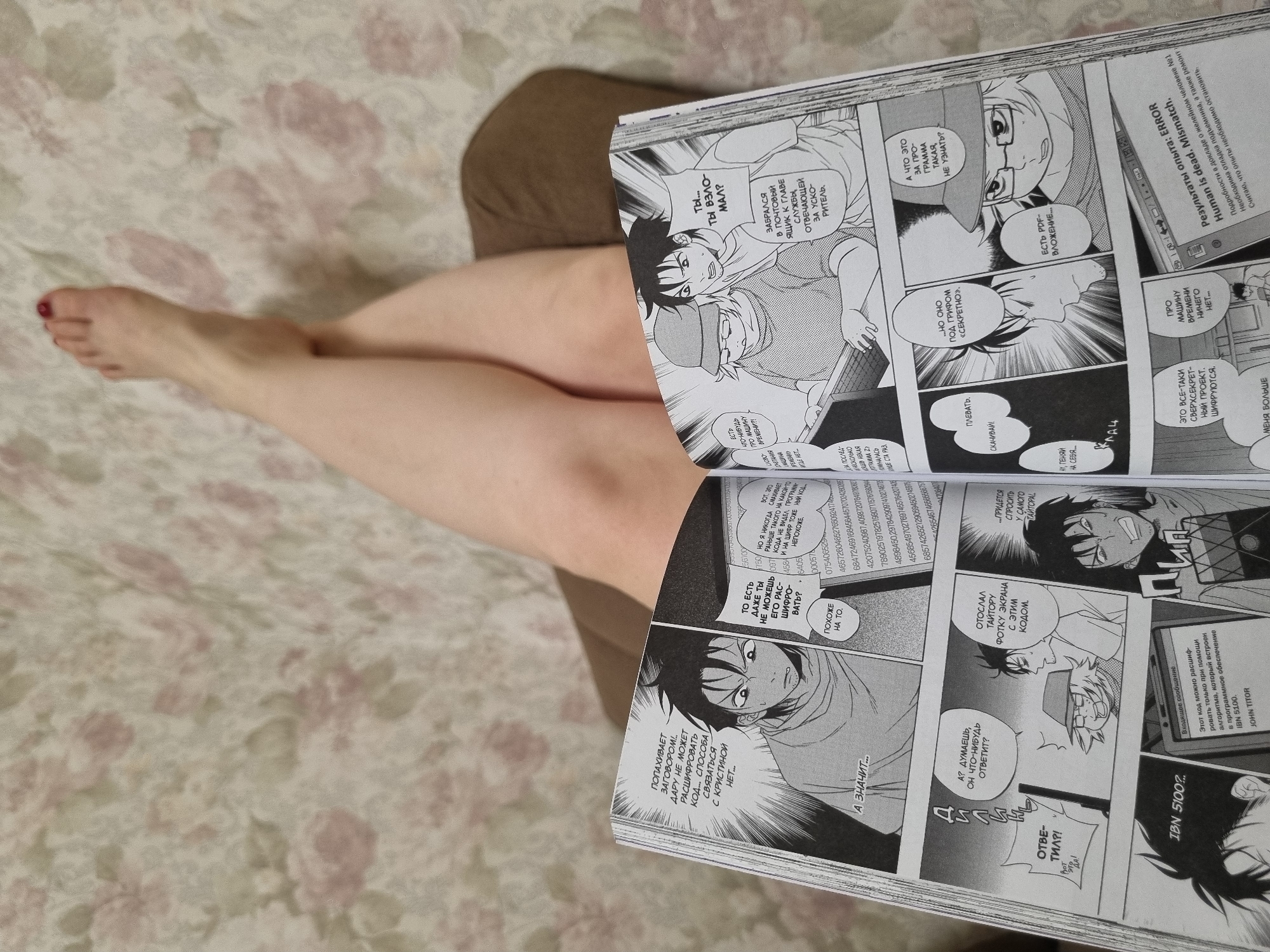 A photograph shows a person sitting on a patterned bedspread, holding open a manga book to display its contents. The book features black-and-white comic panels with Russian text, depicting dialogue and action scenes. The person's legs and one foot are visible, suggesting a casual setting. The scene seems to be indoors, likely at a private residence, given the bedspread and personal items. There's nothing to determine this location is Chaikovsky, Russia.