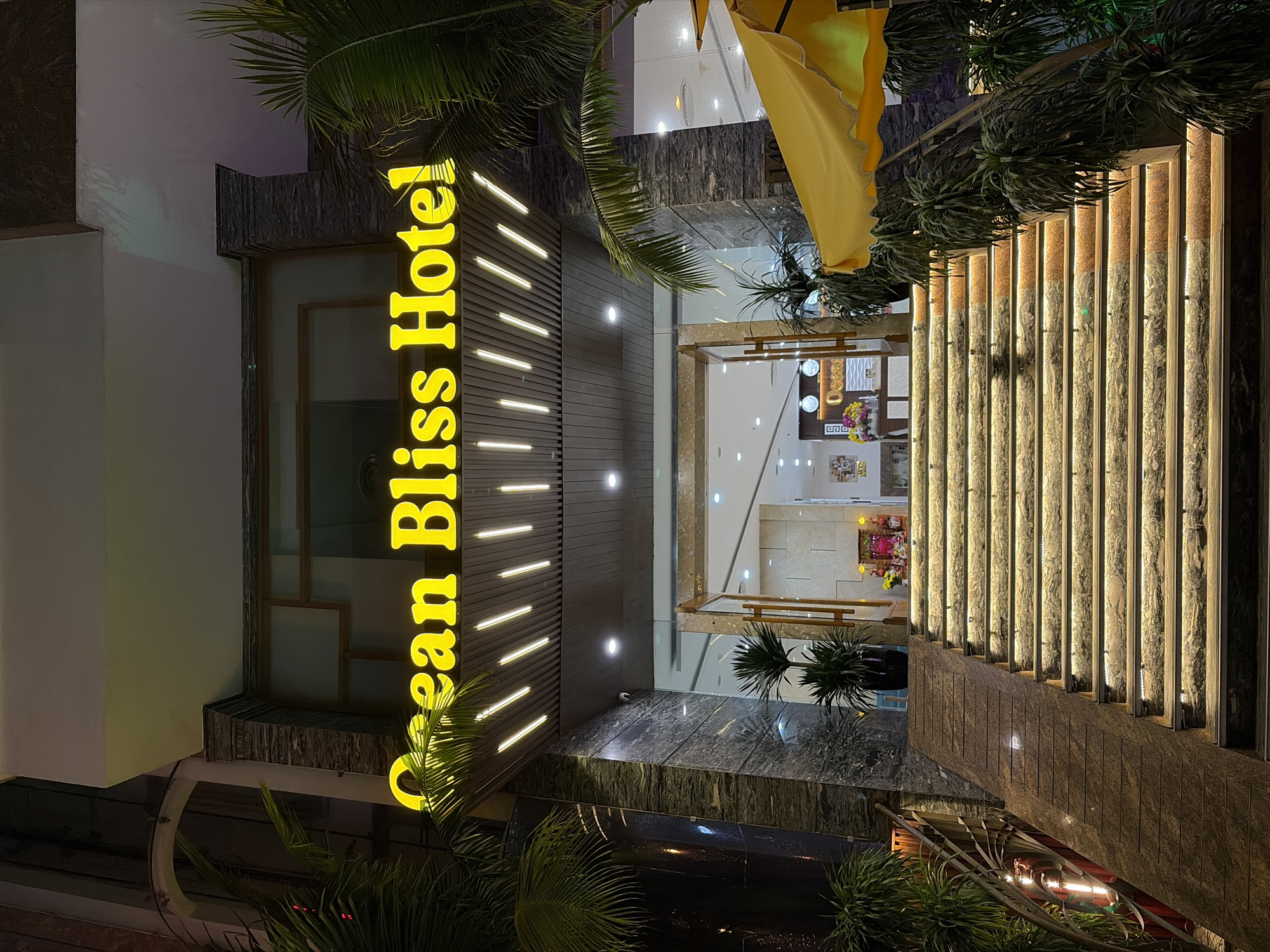 This is a nighttime, eye-level, outdoor, full shot of the entrance to the Ocean Bliss Hotel in Đà Nẵng, Vietnam. The hotel features a bright yellow neon sign spelling out "Ocean Bliss Hotel" above a set of illuminated stairs and a ramp. The entrance is flanked by palm trees and potted plants. The stairs are lit from underneath, creating a warm glow. In the background, through the open entrance, a reception desk is visible with clocks and floral decorations. The facade of the hotel is a mix of dark stone and illuminated panels. A yellow umbrella is visible to the right of the entrance.