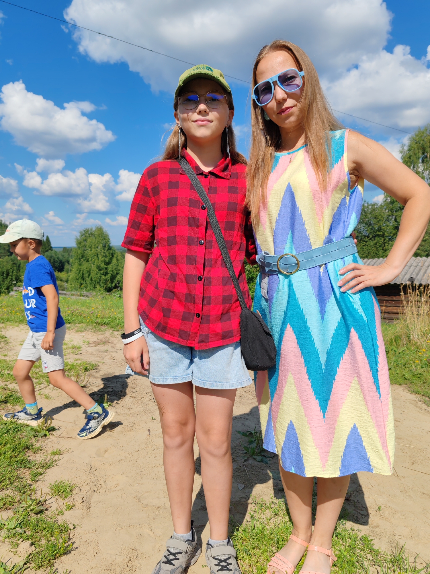 Two females and one male child are standing outdoors on a dirt path. The female in the foreground on the right is wearing a sleeveless, multi-colored, chevron-patterned dress with a blue belt and sunglasses. The female in the foreground on the left is wearing a red and black checkered shirt, denim shorts, and a green baseball cap. The male child, positioned in the background and slightly to the left, is wearing a blue t-shirt, light-colored shorts, and a white baseball cap. He is walking away from the camera. The environment is rural, with green trees and grass visible in the background, under a bright blue sky with white clouds. A wooden structure is partially visible behind the female on the right. The lighting suggests it is daytime.