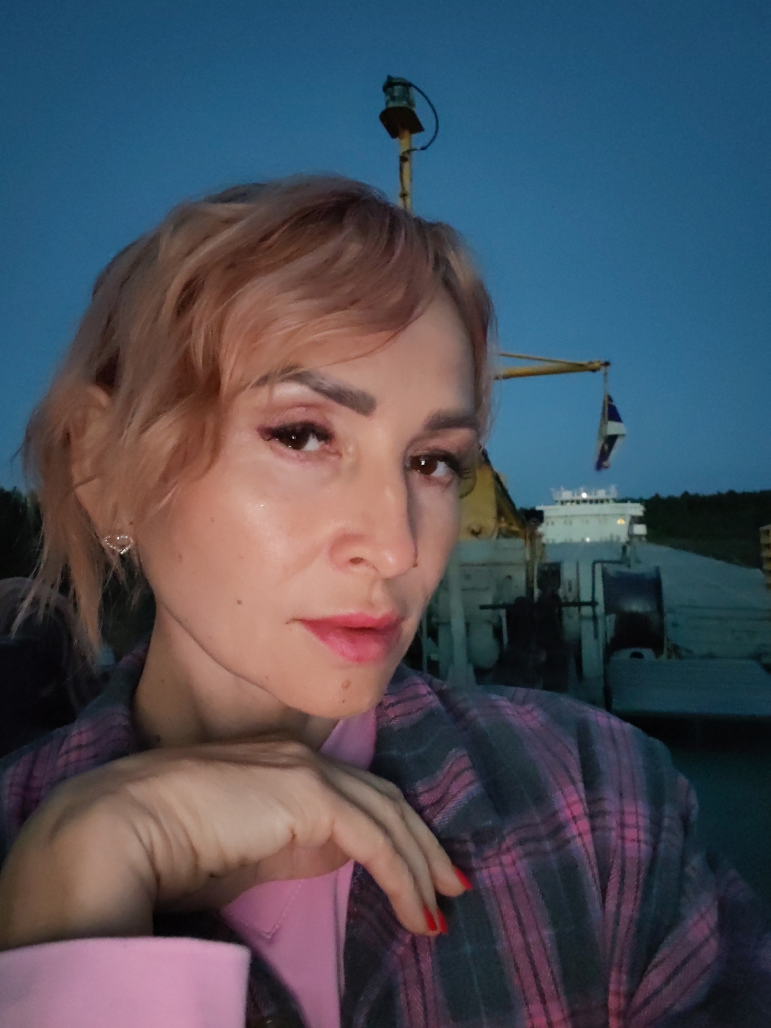 A woman with short, peachy-pink hair, wearing a pink collared shirt and a pink and grey plaid jacket, poses for a selfie at dusk. Her hand rests on her cheek. In the background, a barge is docked on a body of water, with a crane and a flag visible. The sky is a deep blue, indicating the time of day. The location is Annensky Most, Russia.