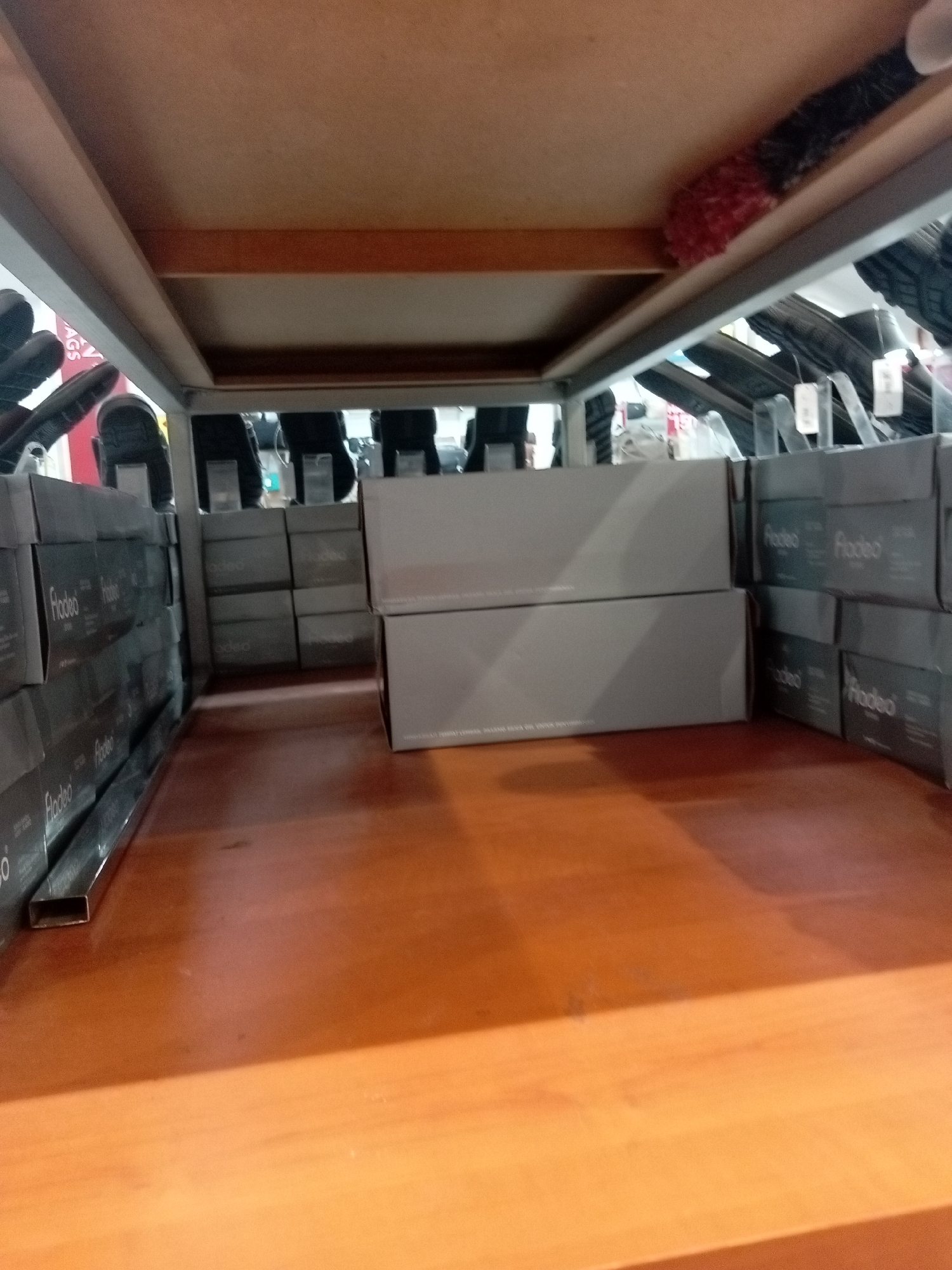The image depicts the interior of a retail store, likely a shoe store, in Batam, Indonesia. The focus is on shelves stocked with shoe boxes. The shelves are made of a light-colored material, possibly wood or laminate, and are part of a display unit that extends from the foreground to the background.
In the foreground, on the left side, are several rows of dark-colored shoe boxes stacked vertically. These boxes appear to be labeled with white text, possibly a brand name. To the right of these boxes, there are two larger, light gray rectangular boxes stacked on top of each other. The top box has some faint white text on its side.
Further into the background, on the right side, are more shelves with shoe boxes. These boxes are also dark, with white labels that seem to repeat a word. The arrangement suggests a retail display where products are organized for sale.
Above the shelves, a wooden surface is visible, forming the underside of another level of the display unit or the ceiling of the store. Partially visible on this surface is a red, fluffy object, possibly a duster or a decorative item.
The lighting in the store appears to be artificial, creating a well-lit environment. The overall impression is of a clean and organized retail space. The presence of shoe boxes strongly indicates that this is a place where footwear is sold. The specific mention of Batam, Indonesia, provides a geographical context for this retail environment.