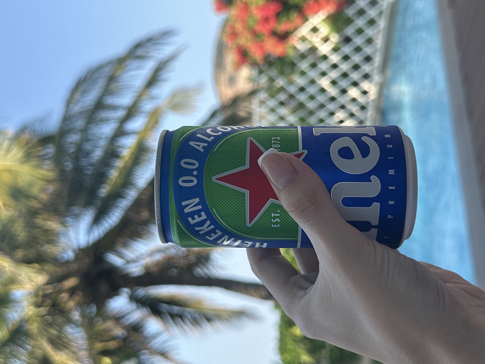 The image shows a hand holding a can of Heineken 0.0 non-alcoholic beer. The background includes a palm tree, a building with red flowers, and a pool with light blue water. The location appears to be outdoors in a sunny area. There are no people visible besides the hand holding the can. The location in the city is Unknown, Unknown.