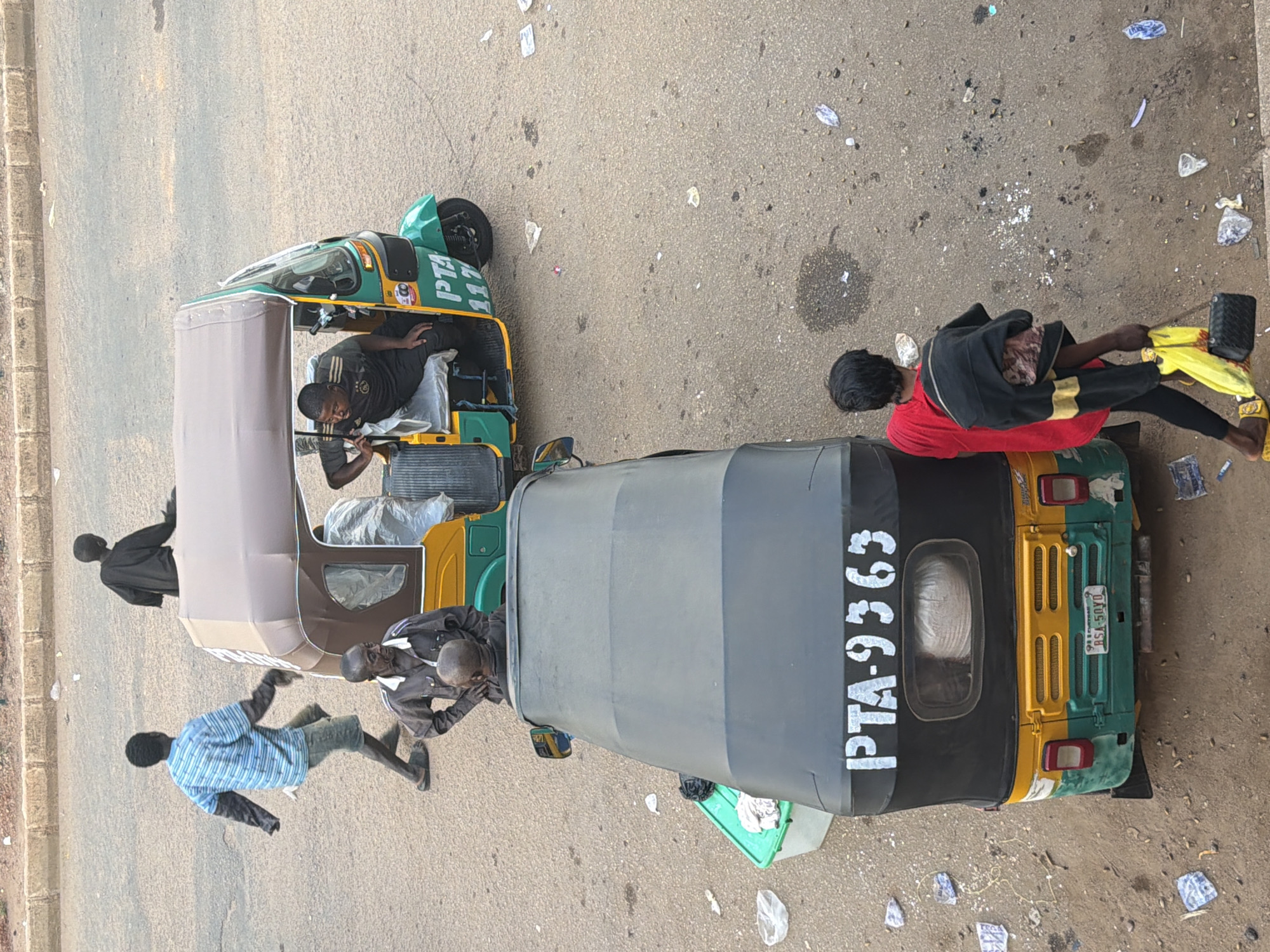 In Bukuru, Nigeria, on a paved road, a scene unfolds with multiple people and three-wheeled vehicles. In the foreground, a black and yellow three-wheeled vehicle, a Keke Napep, is parked. Its license plate reads "RSA-50YO". A person wearing a red shirt and black pants is walking away from the Keke Napep, carrying a black jacket over their shoulders and a yellow bag and a black purse in their hand. Further up the road, a green and yellow Keke Napep is in motion. It has "PTA 117" written on its side. Inside, a person is seated in the driver's position. Another Keke Napep, primarily brown and yellow, is also visible. Two individuals are near this vehicle: one in a dark outfit is standing with hands on hips, and another in a blue and white striped shirt and shorts appears to be dancing or moving energetically. A person in a black outfit is seated in the back of this vehicle. The environment is an asphalt road littered with various debris, including plastic bags and wrappers. The overall impression is a snapshot of daily life and transportation in a Nigerian urban setting.