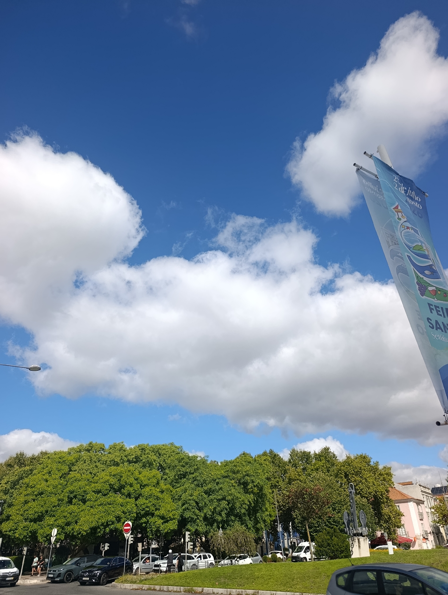 The image depicts a street scene in Setúbal, Portugal, on a sunny day with scattered clouds. A tall banner, likely advertising an event with the text "22 de Julho 2 de agosto" and "FEIRA SAN" visible, is angled against the bright blue sky. In the foreground, a grassy area slopes down towards a street where several cars are parked and driving. Among the parked vehicles are a dark grey SUV and a black sedan. People can be seen standing on a sidewalk and near the cars. Behind the street and cars, a dense line of lush green trees forms a verdant backdrop. In the distance, buildings with pink and beige facades are visible, along with a street light. The overall impression is one of a clear, bright day in an urban environment with abundant greenery.