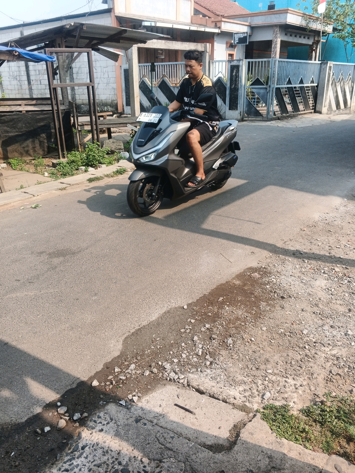 A young man wearing a black t-shirt with yellow accents and black shorts rides a gray scooter down a street in Kadu, Indonesia. He is looking forward and appears to be smoking. The scooter has a license plate that reads "B 6328 JSD". The street is paved with asphalt and has a gravel shoulder on the right side. There are houses and fences visible on the left and right sides of the street. A flag is flying from a pole in front of one of the houses. The scene is illuminated by sunlight, casting shadows on the street.