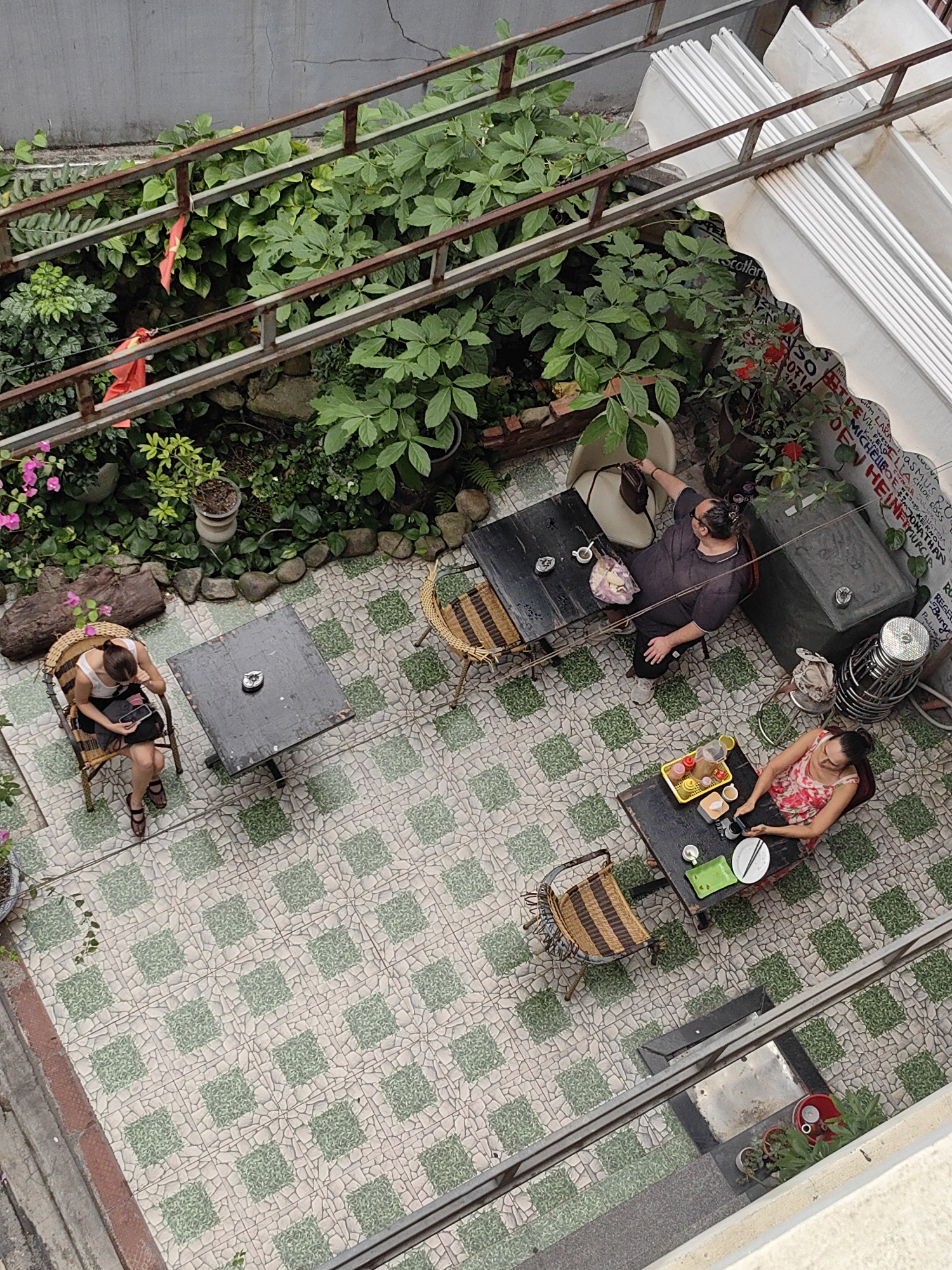 This is an aerial view of an outdoor courtyard cafe located in Bai Chay Ward, Vietnam. The cafe features a patterned tile floor and is surrounded by lush greenery, including plants and flowers. Three people are seated at separate tables. One woman is sitting at a table in the left of the frame, engrossed in her phone. Another person sits alone at a table, reaching for a bag placed on an empty chair, and a third woman is at a table set with food and drinks. A red flag is seen at the center top behind the top of the table with the man. The cafe is bordered by metal railings. A wall covered in graffiti is visible in the upper right.